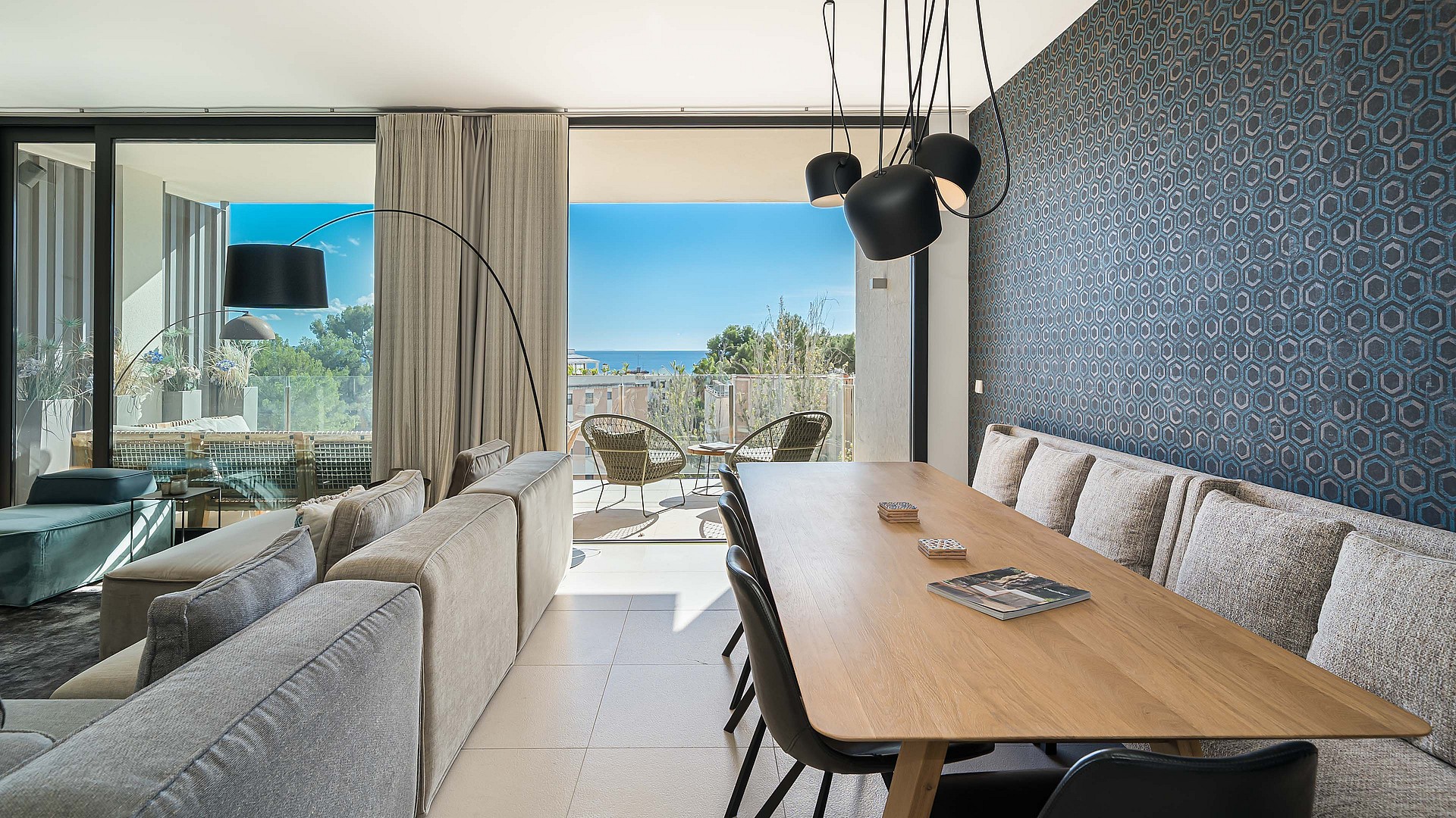 Interior view of an open-plan living and dining room with floor-to-ceiling glass doors opening onto a terrace with a sea view.