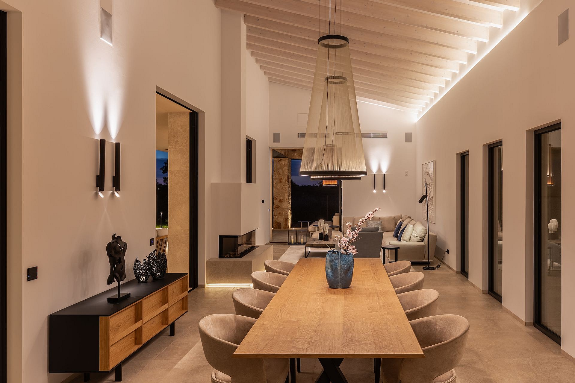 Dining room with a long oak table and curved chairs, featuring a high gabled ceiling with exposed beams and recessed lighting.