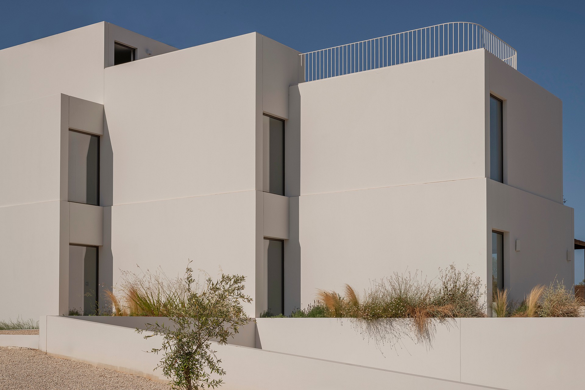 Modern white geometric building exterior featuring sharp vertical lines, small windows, and native grasses in the foreground.