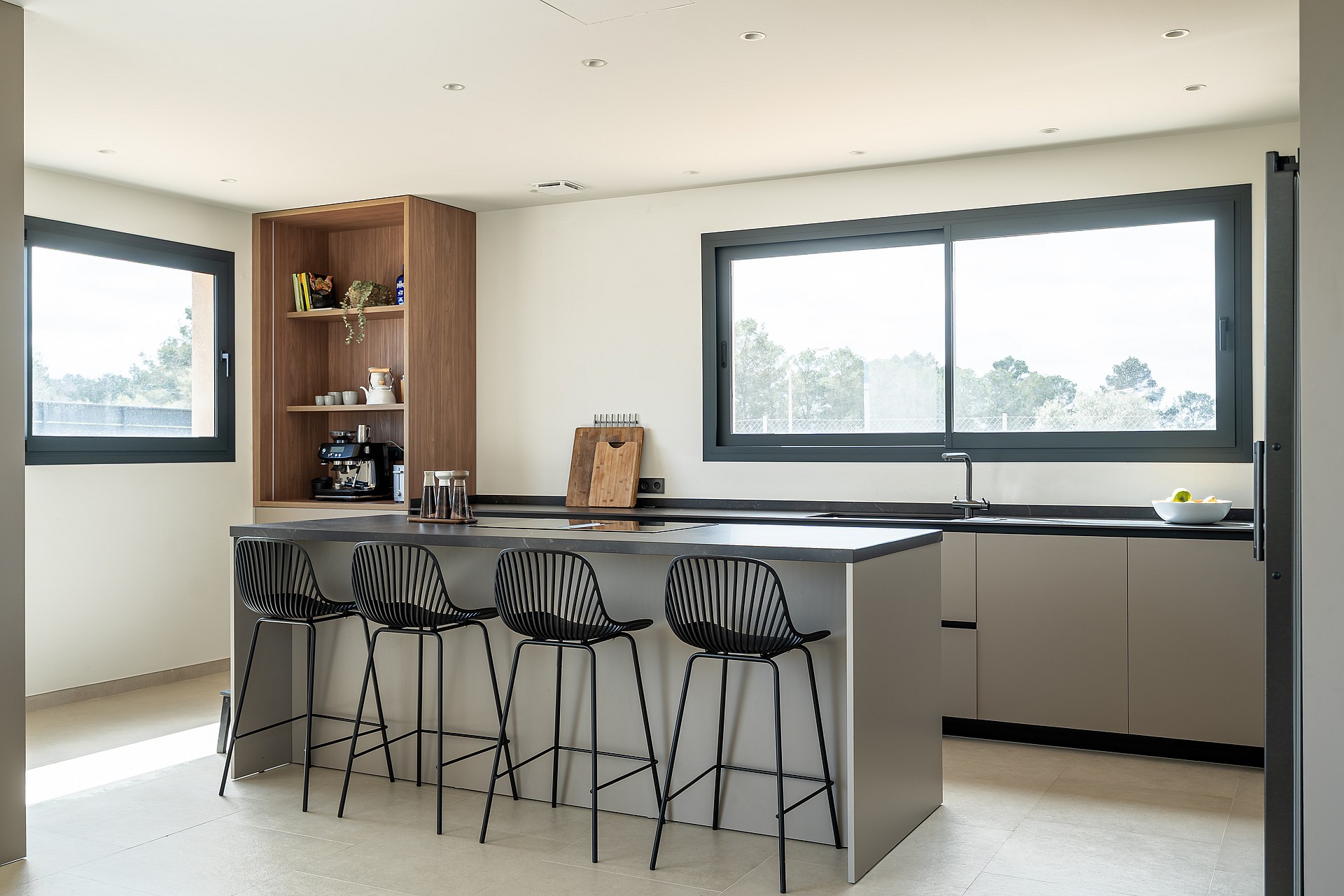 A modern kitchen with a grey island, black stools, and wooden open shelving, illuminated by natural light from windows.