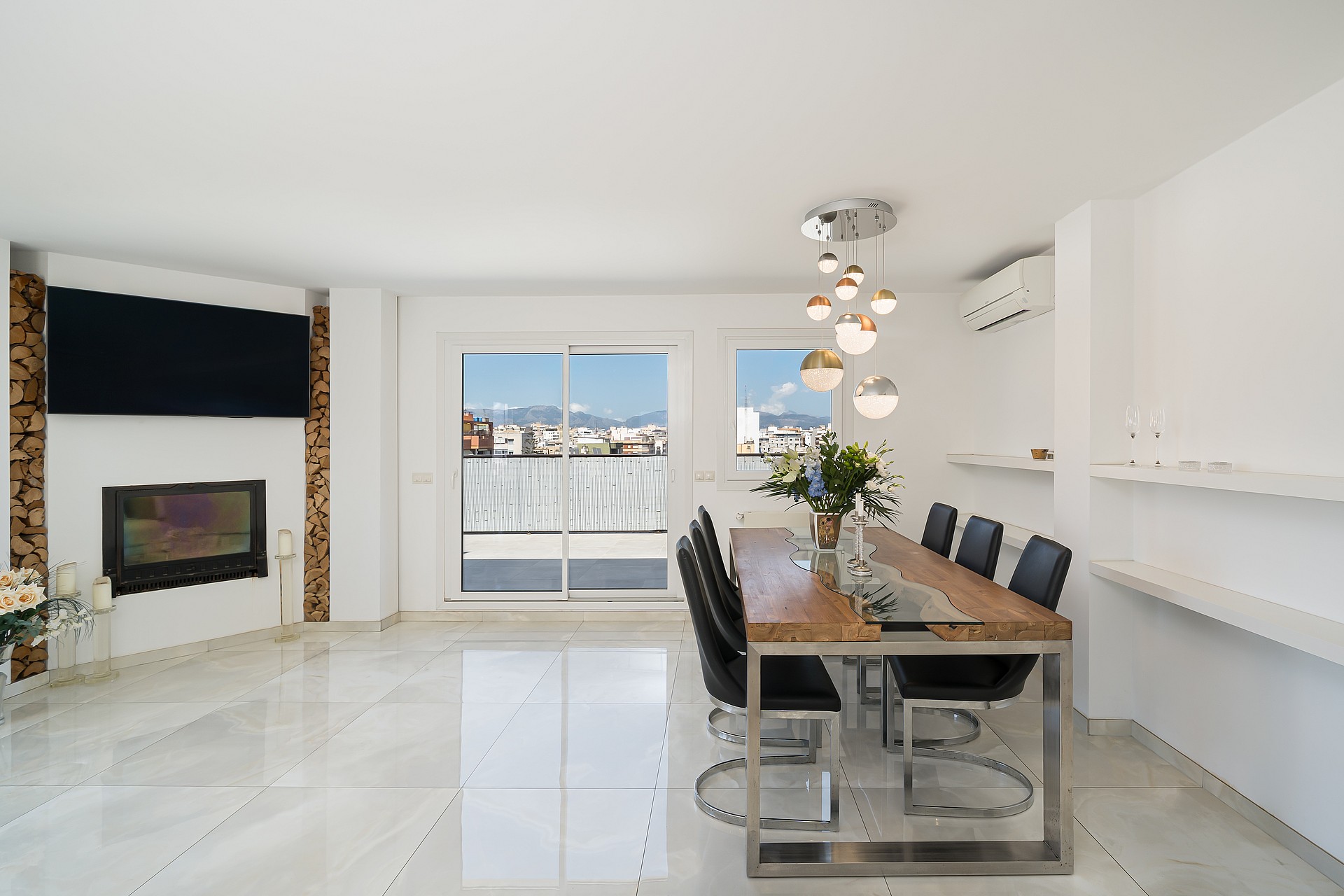 Dining room with a wooden table, black chairs, and a fireplace with a mounted television, featuring city views through glass doors.