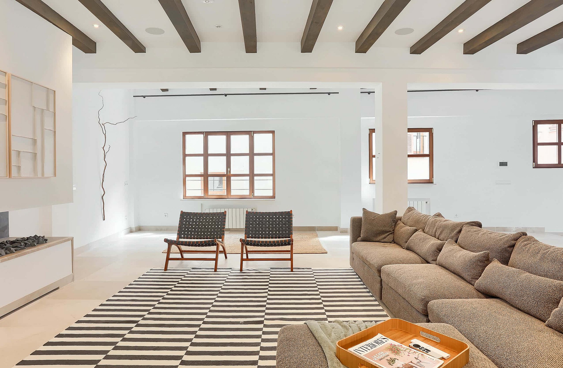 A bright, open living room with dark wooden ceiling beams, white walls, a large modular sofa, and two leather chairs.
