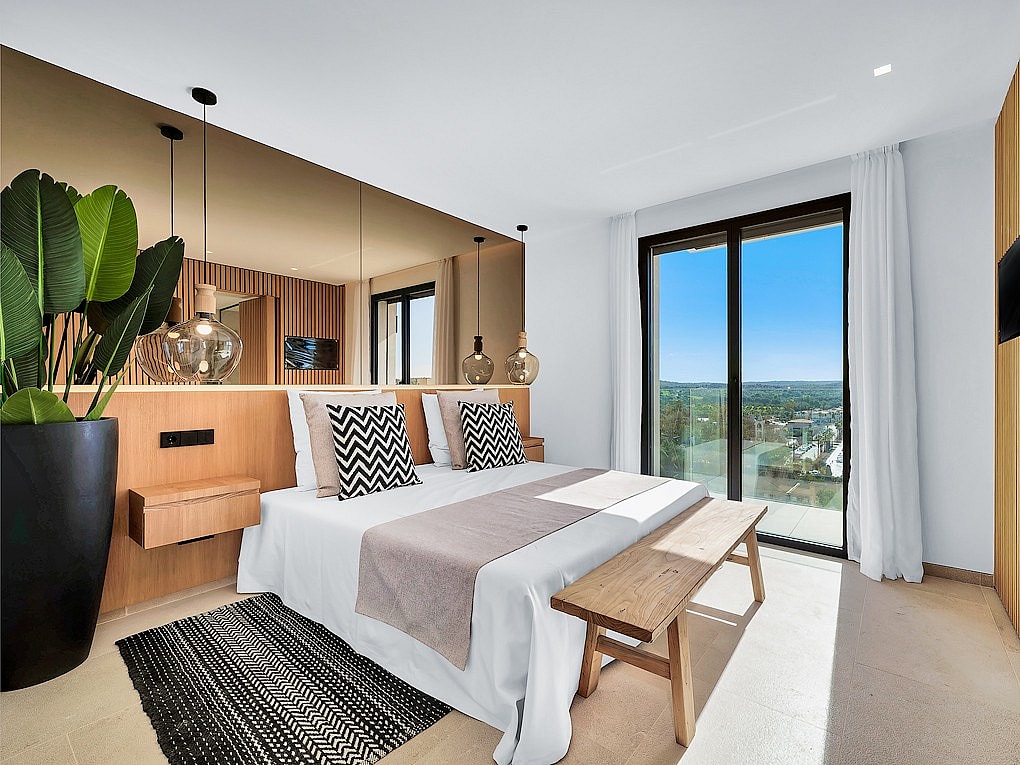 A modern bedroom featuring a bed with a wooden headboard, a large mirror, a wooden bench, and a floor-to-ceiling glass door.
