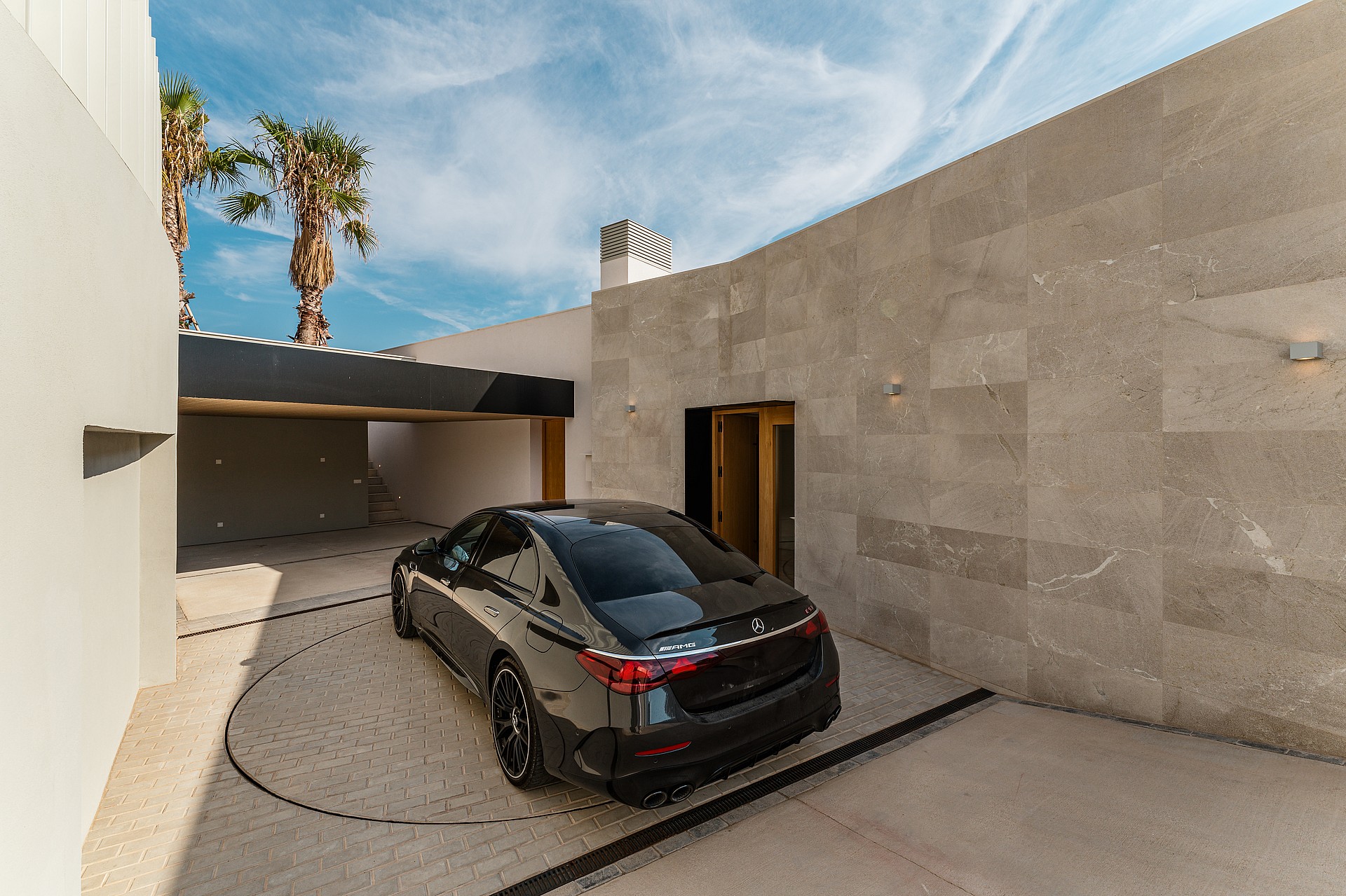 Modern outdoor courtyard featuring a stone-paved turntable for a car, shaded carport, and smooth beige walls under a blue sky.