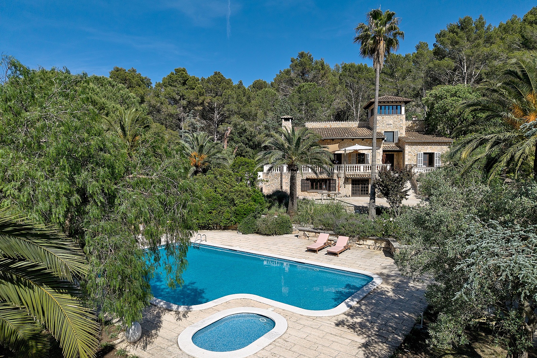 Exterior view of a traditional stone finca surrounded by trees, featuring a rectangular swimming pool and a separate plunge pool on a stone patio.