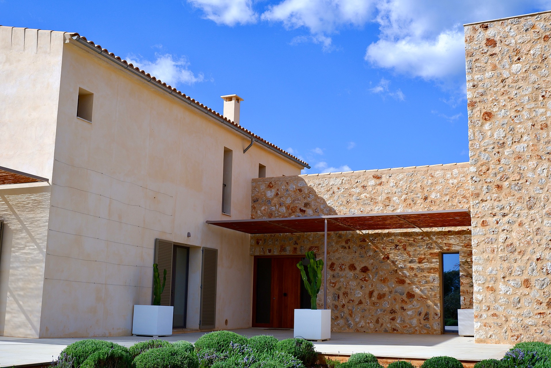 Exterior view of a stone and plaster finca featuring a terrace area with a wooden pergola, potted plants, and clear blue sky.
