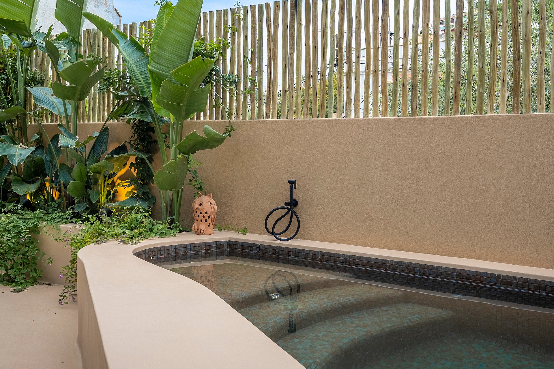 A small plunge pool with dark mosaic tiling, set into a beige rendered wall with a bamboo privacy screen and tropical plants.