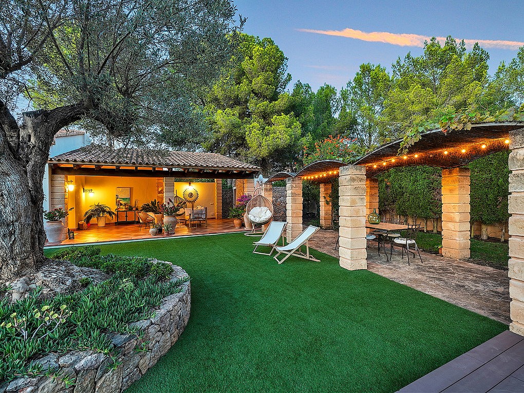 A landscaped garden with a large ancient olive tree, stone-pillared pergola with string lights, and a shaded outdoor room.