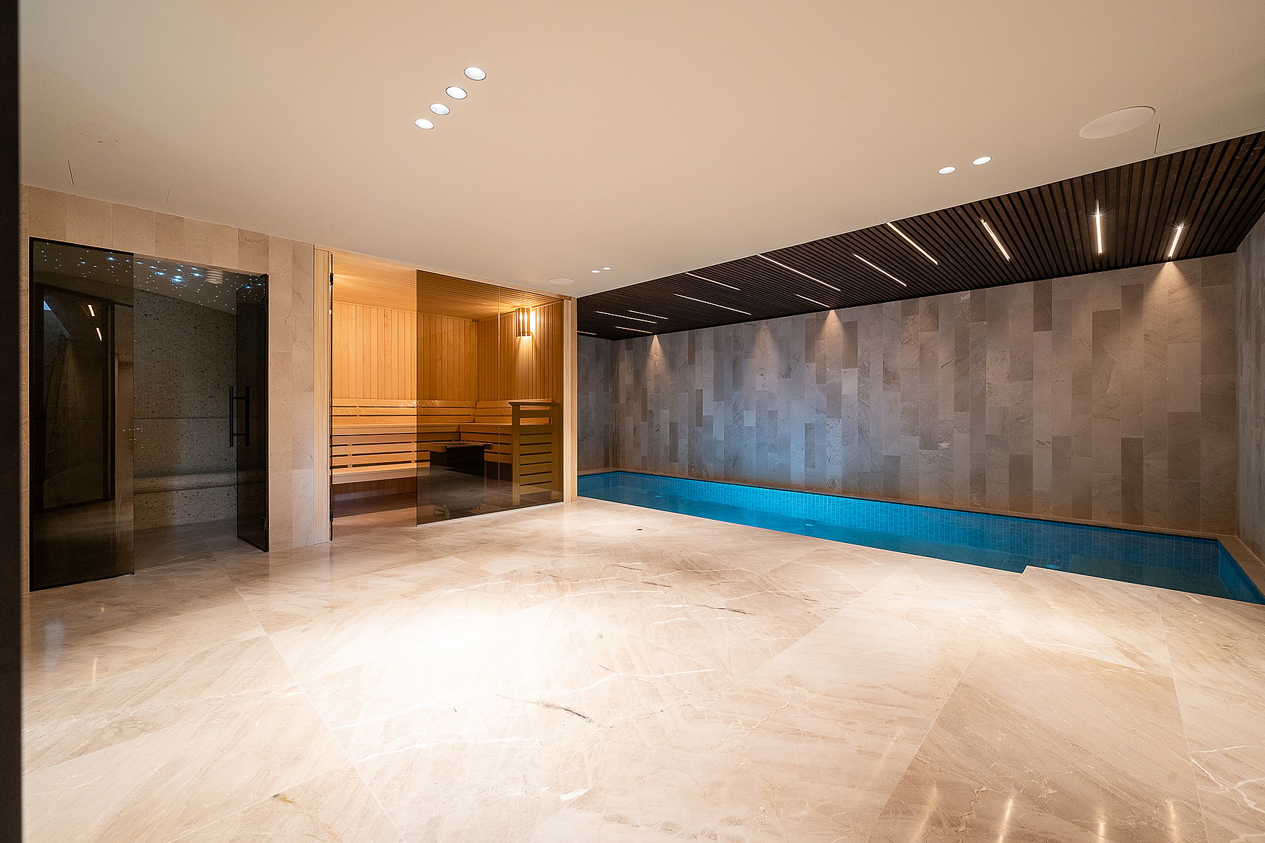 Indoor spa area featuring a tiled lap pool, sauna with glass door, polished stone flooring, and recessed ceiling lighting.