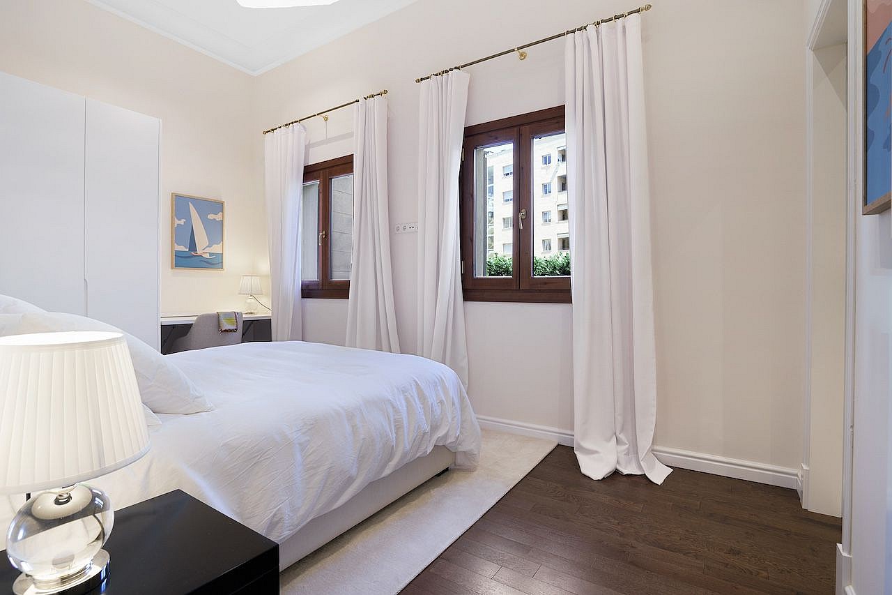 A minimalist bedroom featuring dark hardwood flooring, white walls, white bedding, and two windows with sheer curtains.