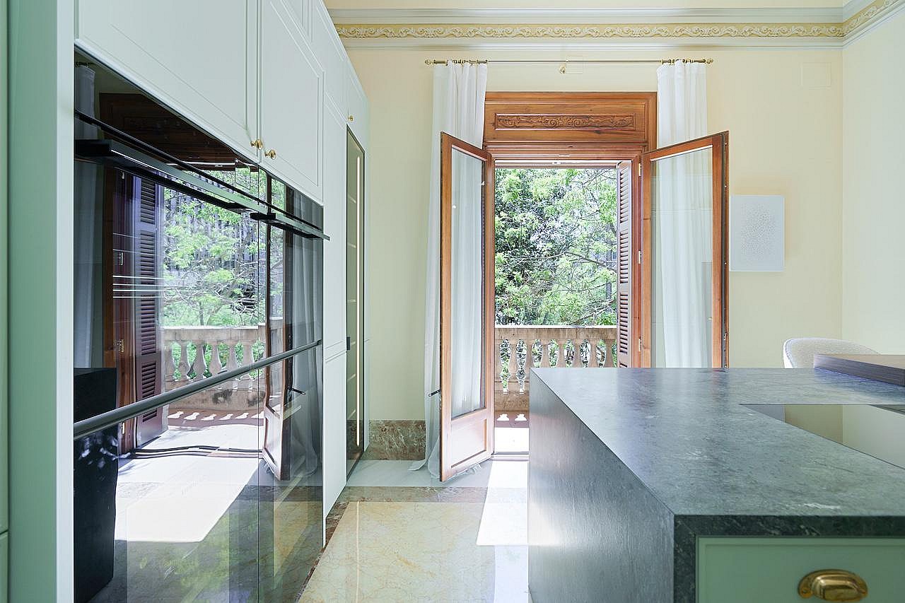 Modern kitchen featuring a dark stone island, sleek white cabinetry, and French doors opening to a stone-railed balcony.