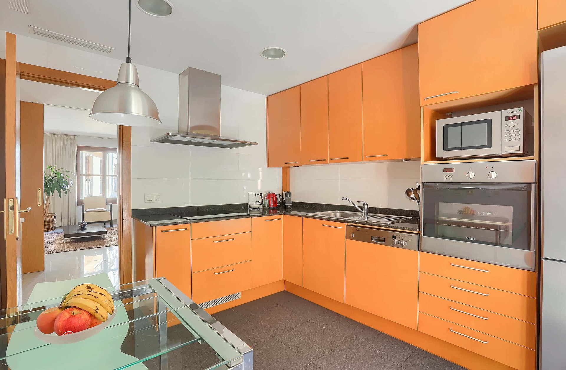 An L-shaped kitchen featuring bright orange flat-panel cabinets, black stone countertops, and a glass-topped dining table.