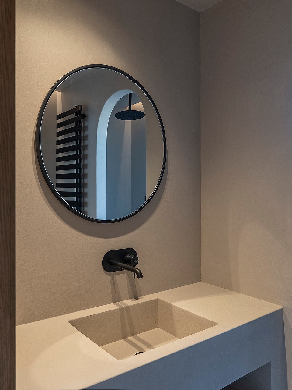 A minimalist integrated stone sink and wall-mounted black faucet beneath a circular mirror reflecting a shower area.