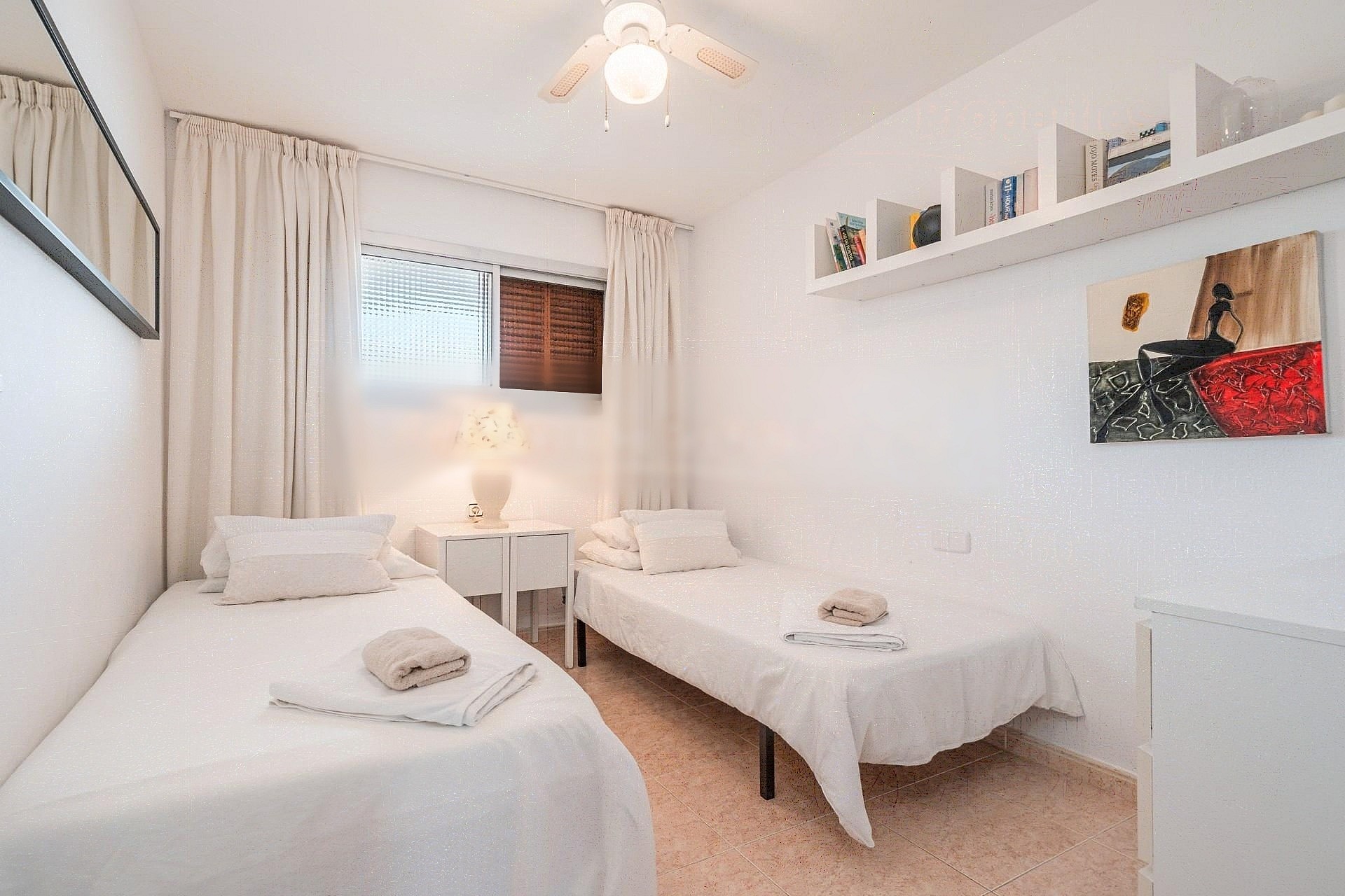 A bright guest bedroom featuring two single beds, light-toned textiles, a white wall-mounted shelf, and a minimalist aesthetic.