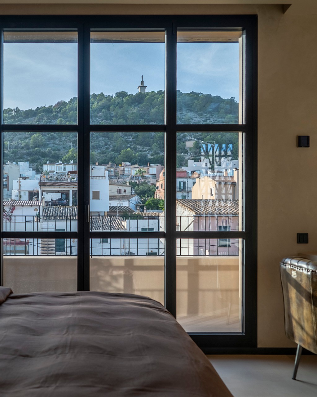 A bedroom featuring a large grid-style window overlooking a Mediterranean town and green hillside.