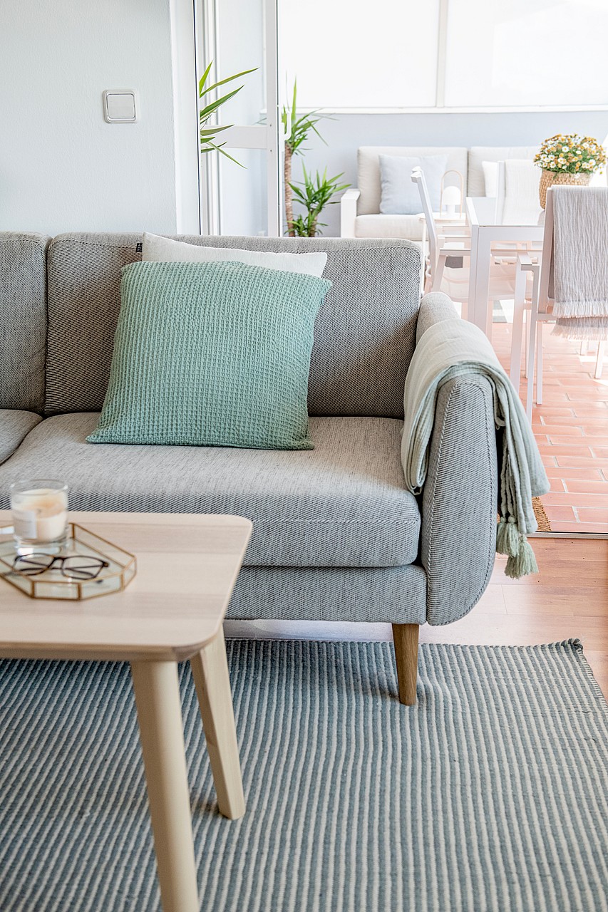 Close-up of a grey woven sofa with a mint green cushion and a light wood coffee table on a striped rug, leading to a terrace.
