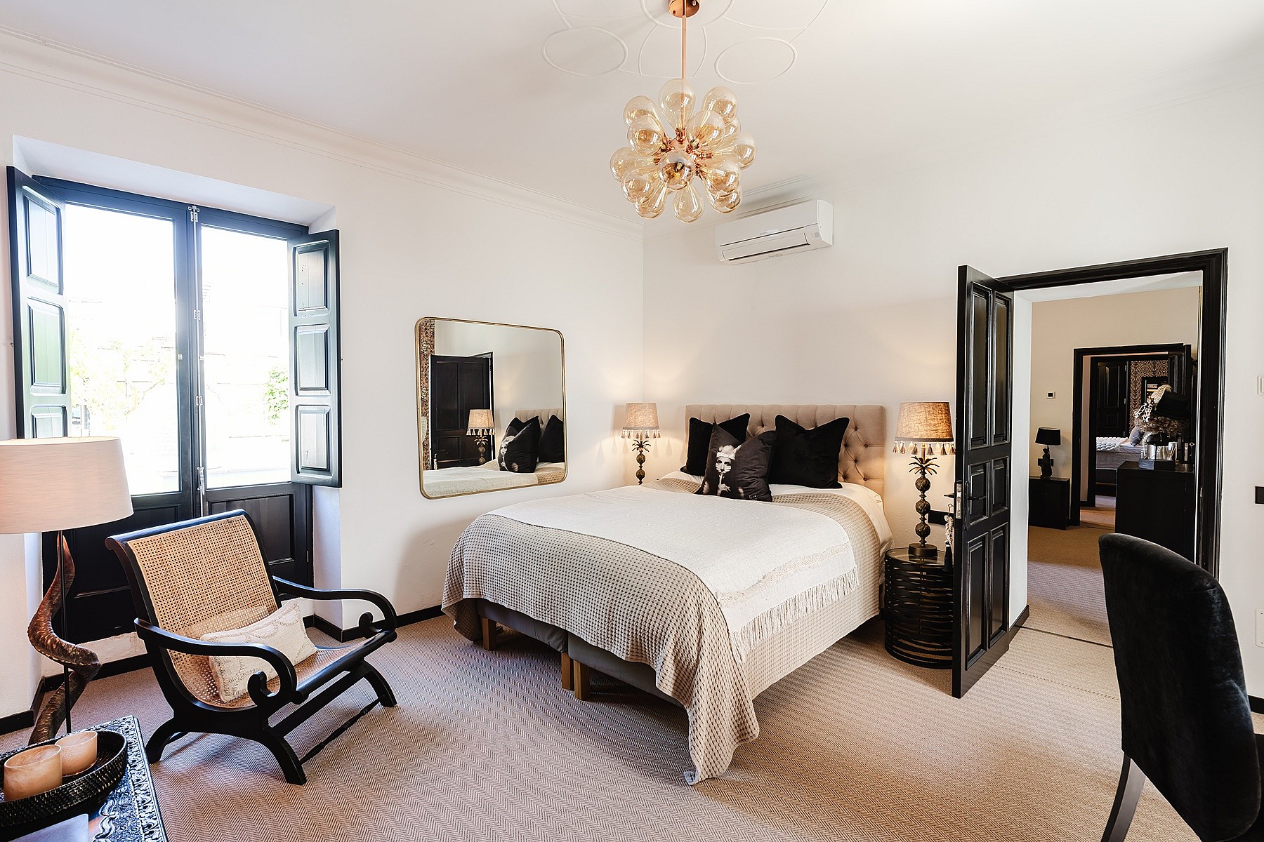 A spacious bedroom featuring a king-sized bed with dark accent pillows, a woven armchair, and an open doorway leading to another room, illuminated by a chandelier.