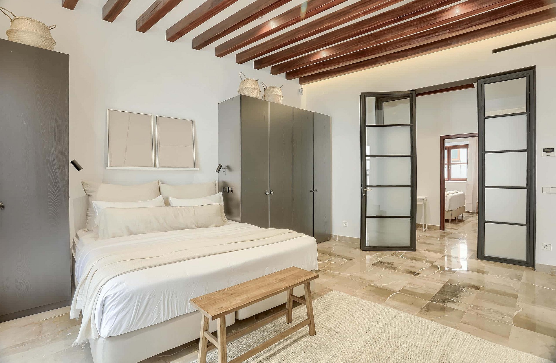 A spacious bedroom featuring exposed dark timber ceiling beams, white walls, stone flooring, and dark metal-framed glass doors.