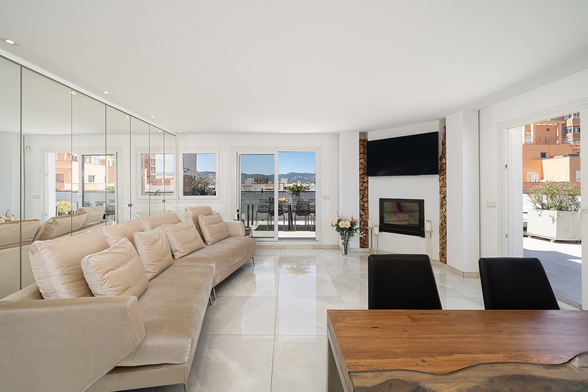 A bright, spacious living room with polished white floors, a long beige sofa, floor-to-ceiling mirrored walls, and a contemporary fireplace.