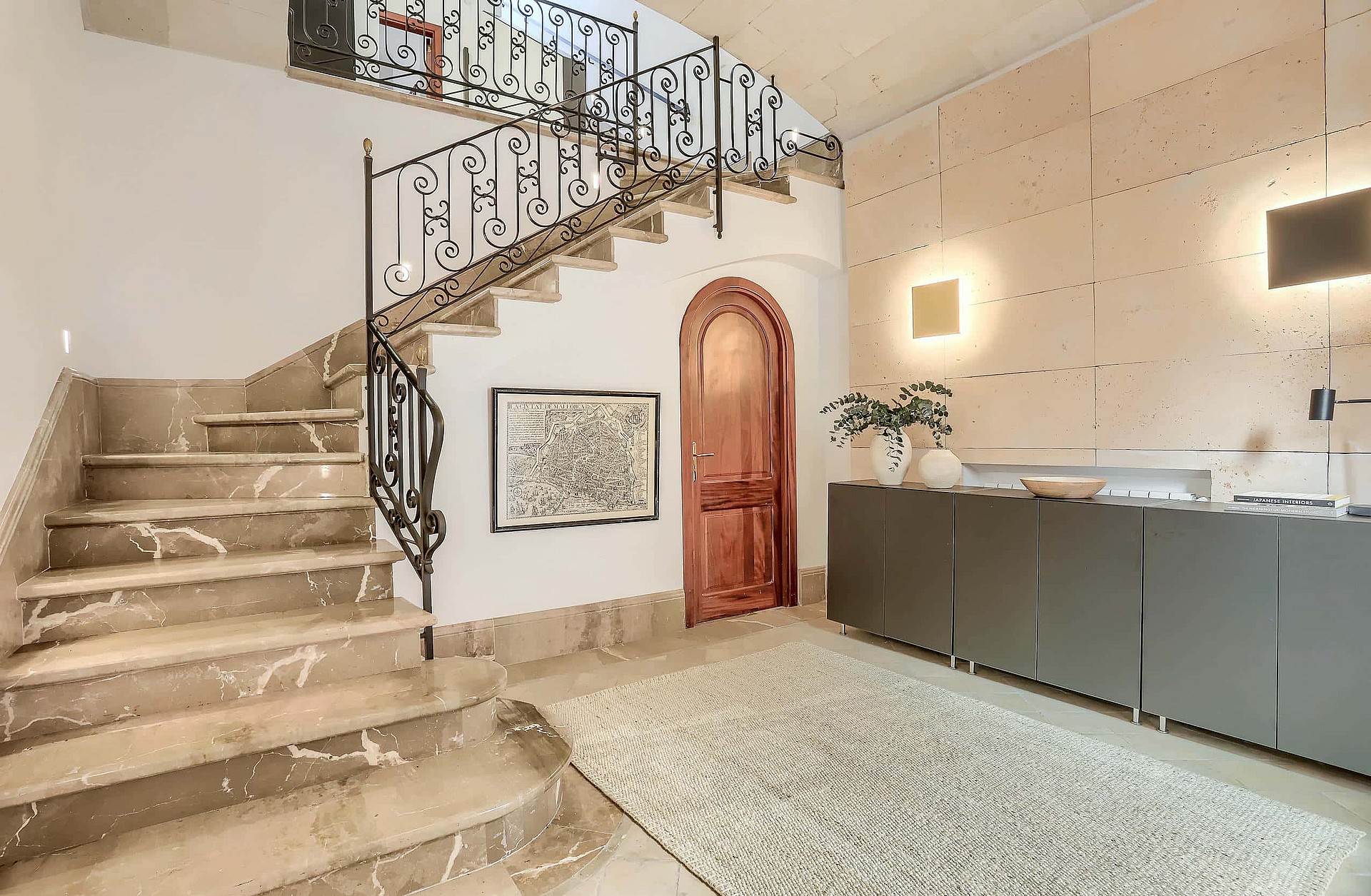 An interior shot of an entrance hall featuring a wide marble staircase with a wrought-iron railing, a curved wooden door, and a textured stone feature wall.