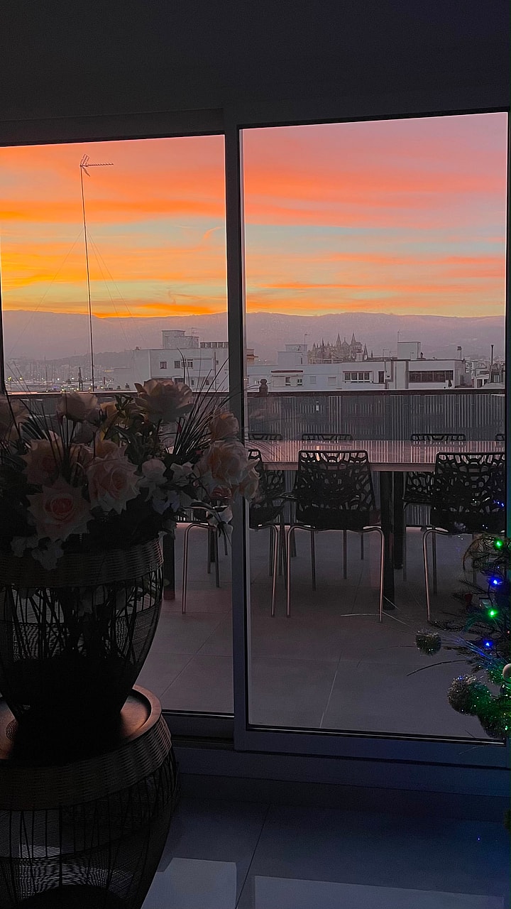 View through a glass door showing an urban skyline at sunset, with a silhouette of an outdoor dining area on the terrace.