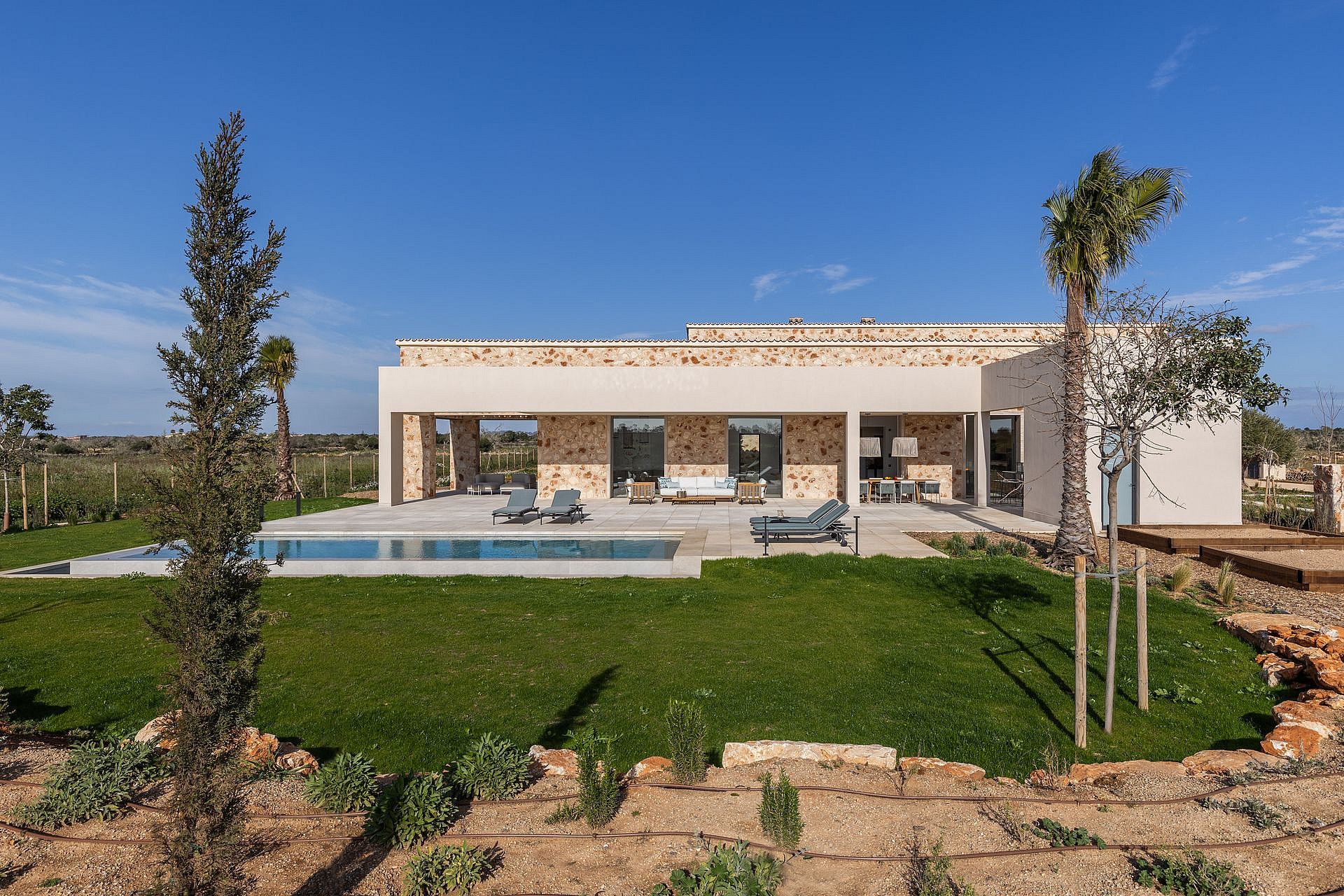 Exterior view of a modern stone finca in Santanyí featuring a rectangular swimming pool, manicured lawn, and sun loungers.