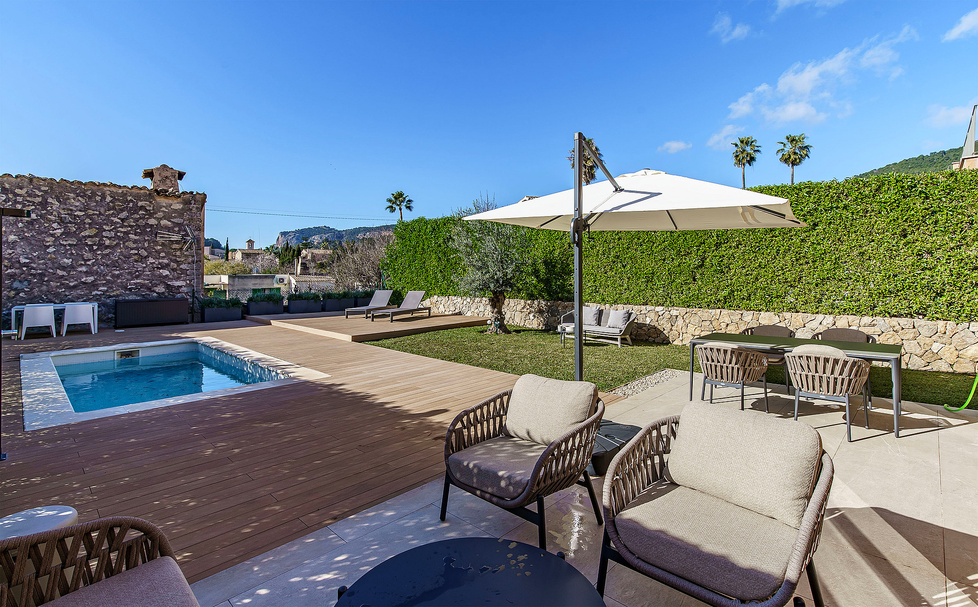 A backyard garden with a small swimming pool, wooden decking, a dining set under an umbrella, and woven outdoor lounge chairs.