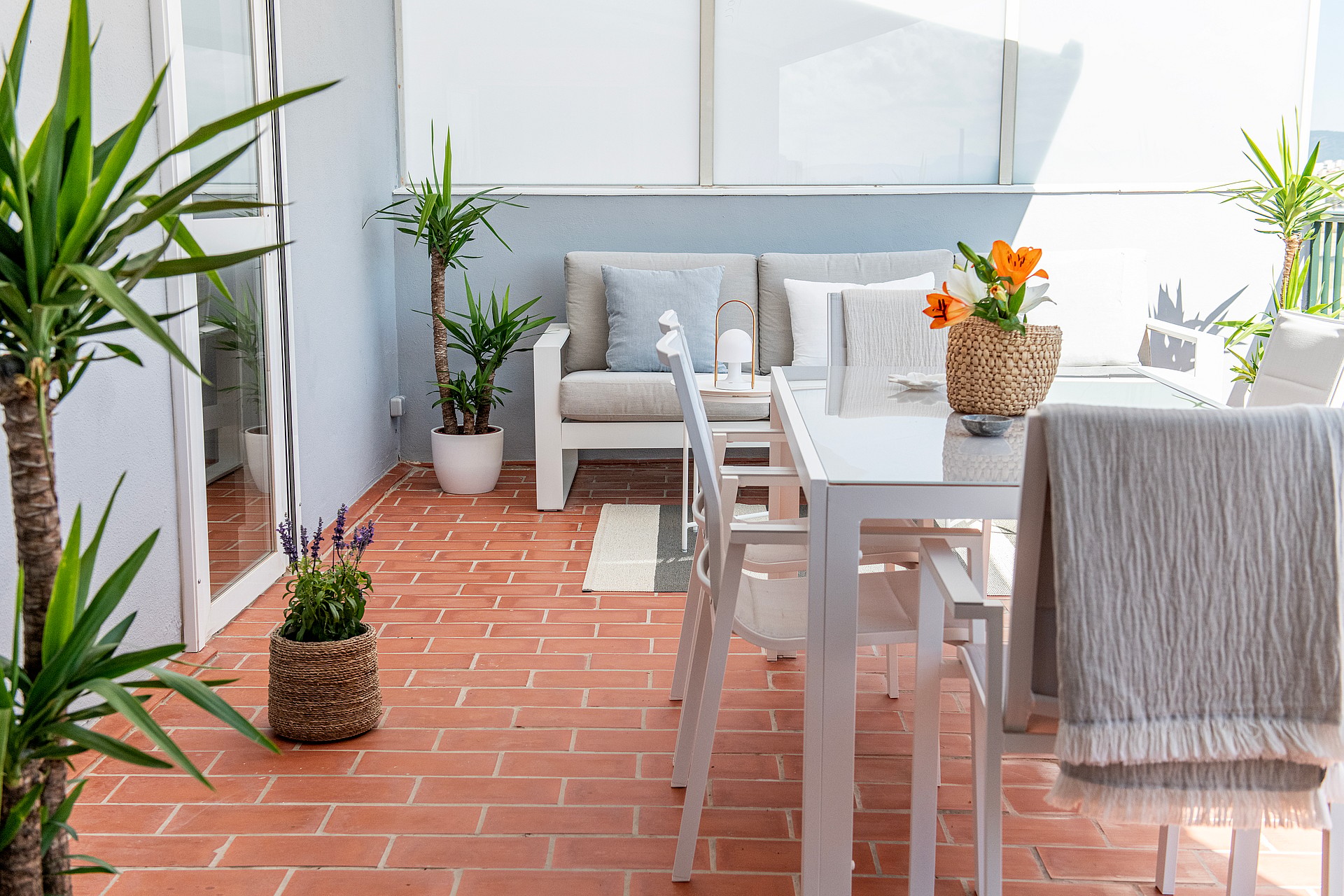 An outdoor terrace with terracotta tile flooring, a white dining table with matching chairs, and potted yucca plants.