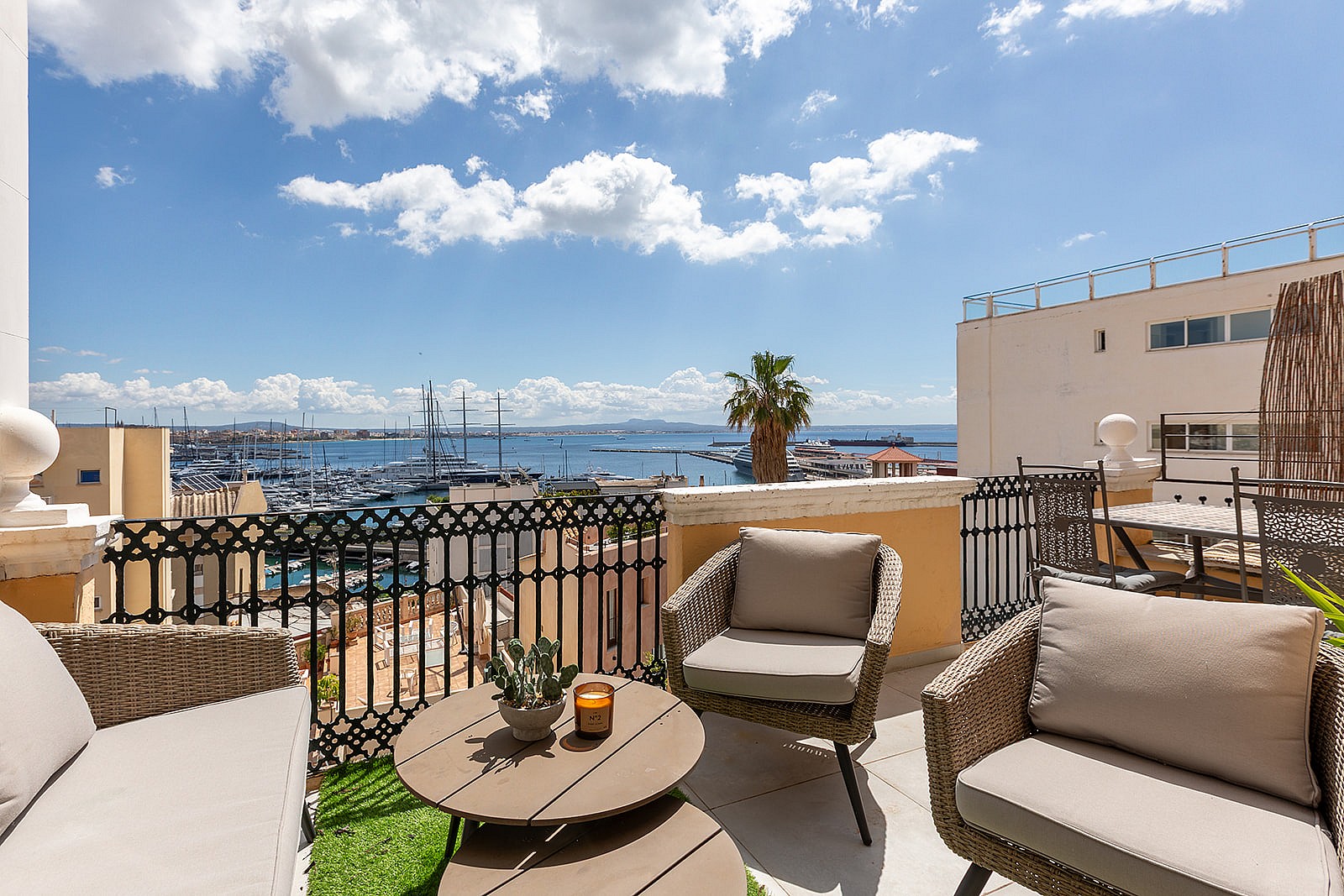 Outdoor terrace with woven armchairs, a round wooden table, and iron railings overlooking a marina and sea under blue sky.