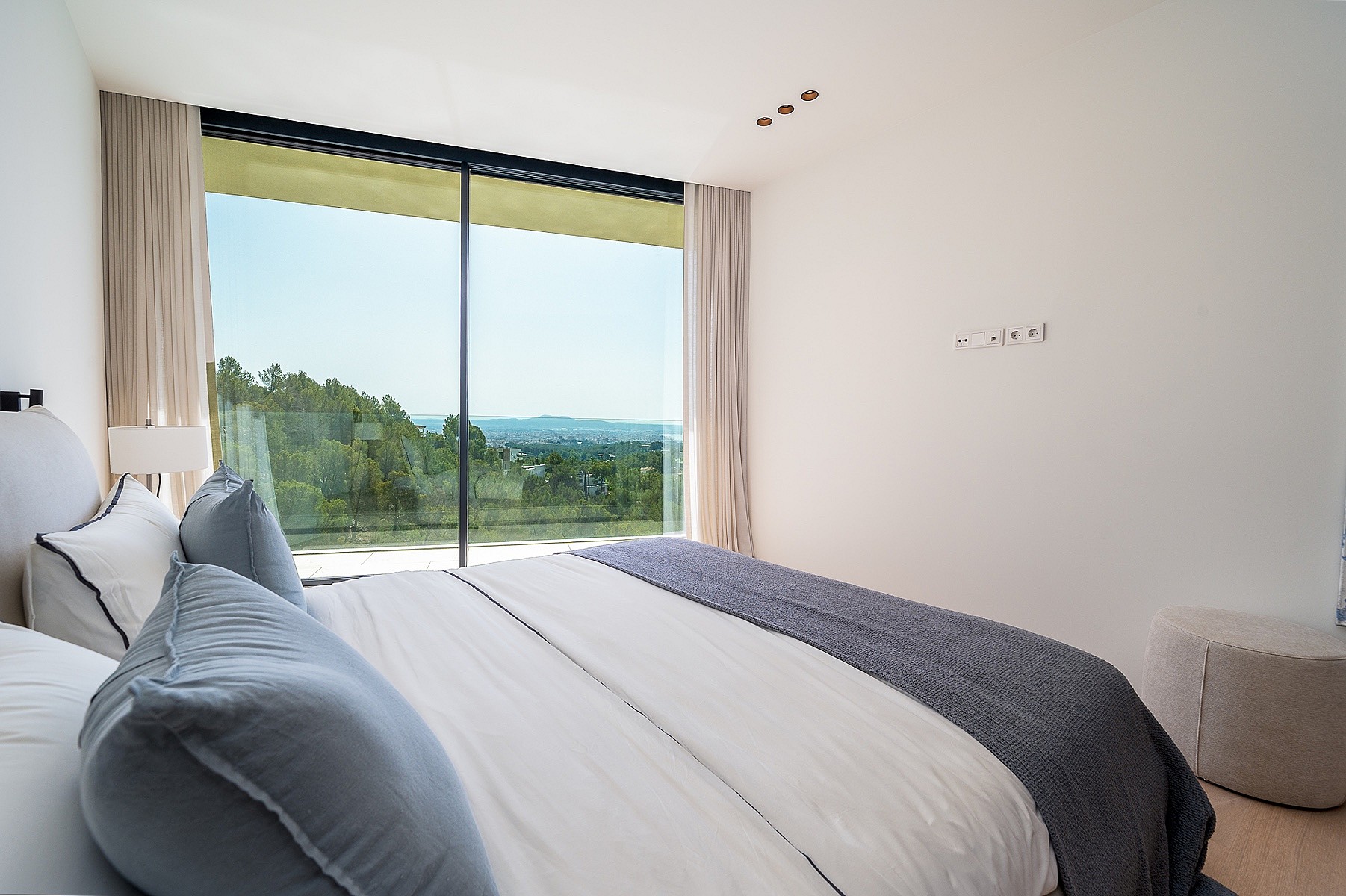 Modern bedroom with a large bed, floor-to-ceiling sliding glass doors, and a distant sea view framed by pine trees.