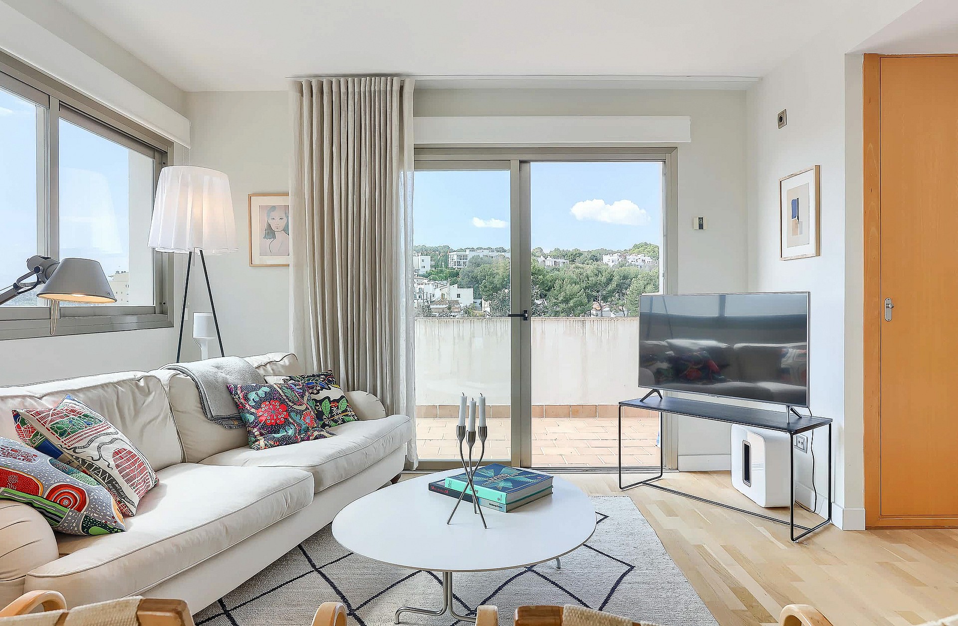 A bright living room with a beige sofa, patterned pillows, round coffee table, and large glass doors opening to a terrace.