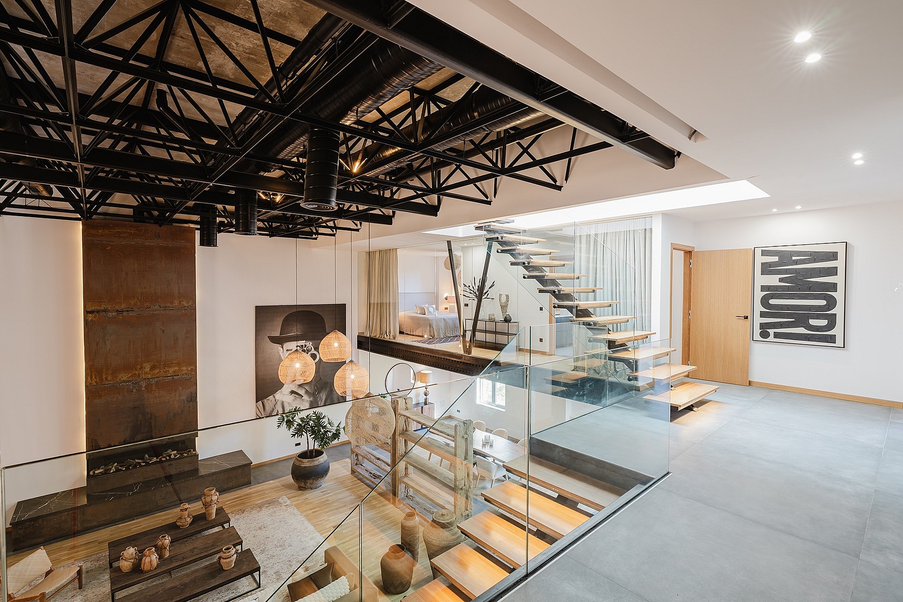 High-angle view of an open-plan apartment featuring a black exposed ceiling truss, glass-railed staircase, and mezzanine level.