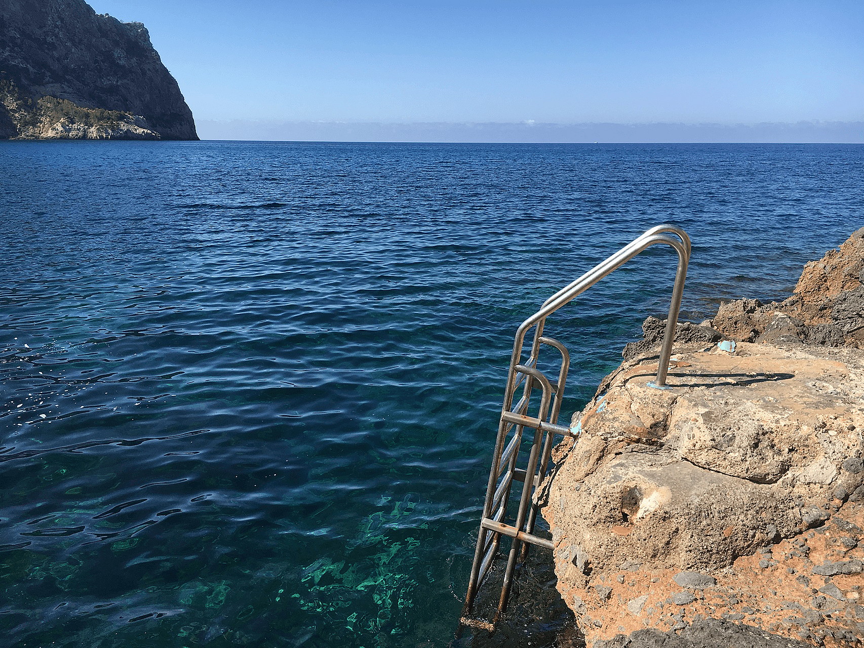 A stainless steel swim ladder attached to natural rocky coastline leading into deep blue Mediterranean sea waters.