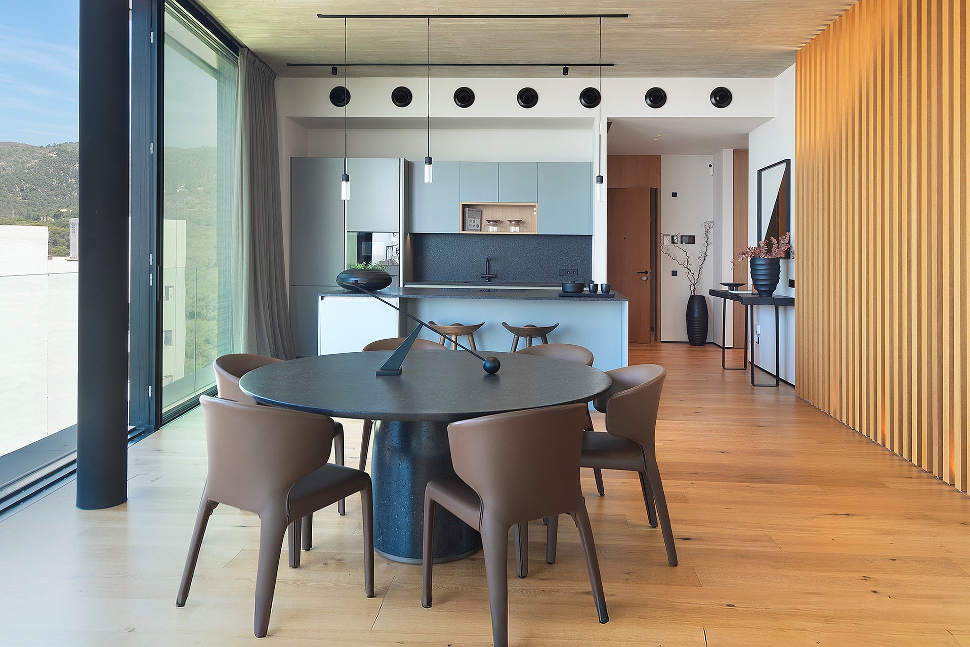 Modern open-plan kitchen with a dark circular dining table, leather chairs, and wooden slat wall paneling, featuring floor-to-ceiling sliding glass doors.