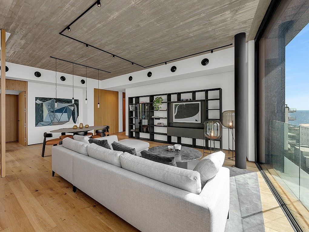 Modern open-plan living room with light oak flooring, concrete ceiling, track lighting, a large light gray sofa, and full-height glass walls overlooking the sea.