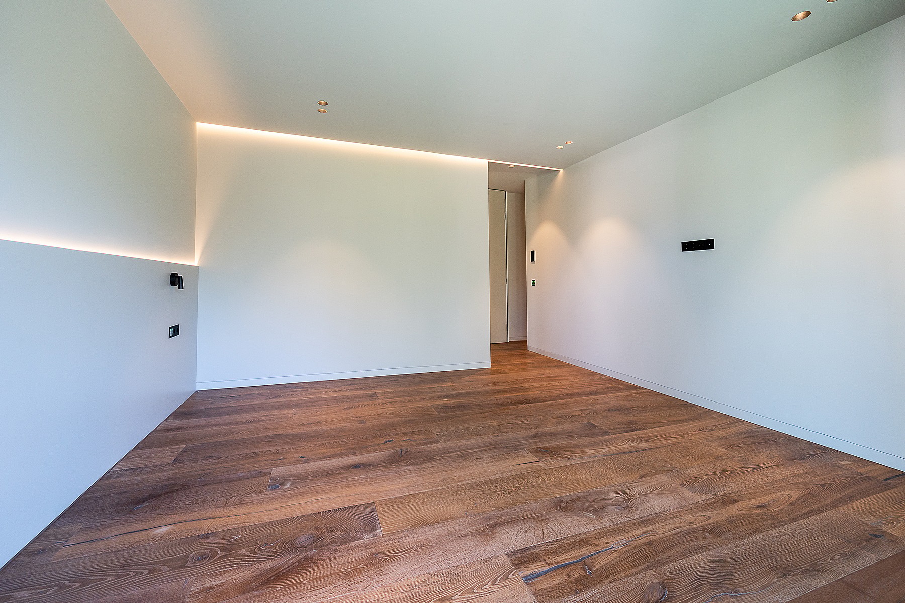 An unfurnished minimalist bedroom featuring white walls, warm LED cove lighting, and wide-plank oak wood floors.