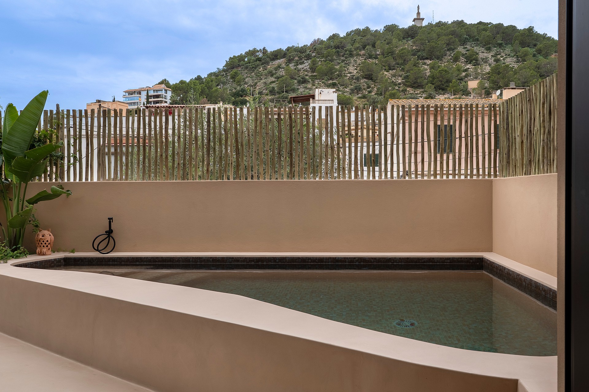 A small triangular plunge pool with beige rendered walls and a vertical bamboo privacy screen, set against a verdant hill.