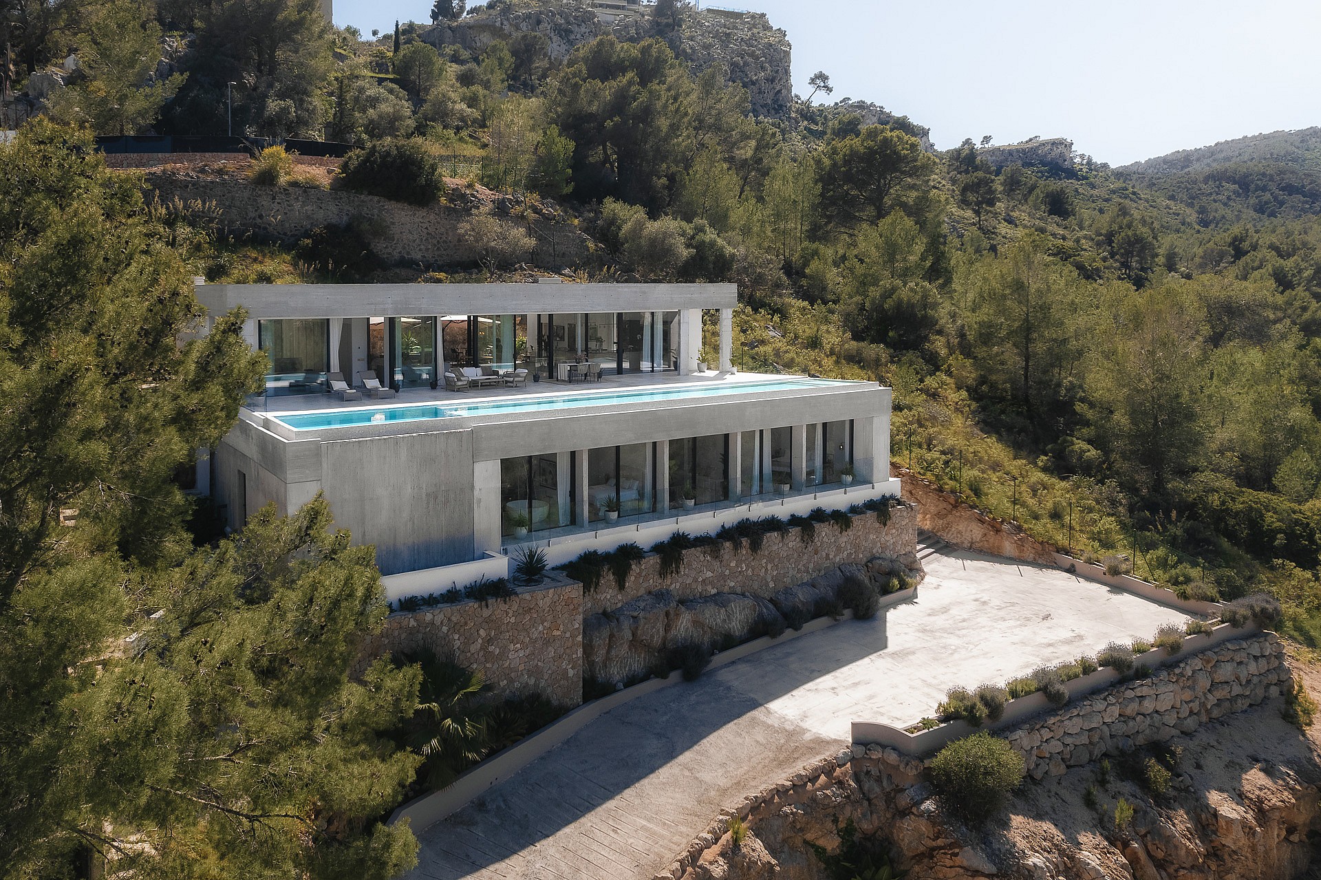 Aerial perspective of a multi-level modern villa featuring an infinity pool, stone walls, and glass facades in Son Vida.