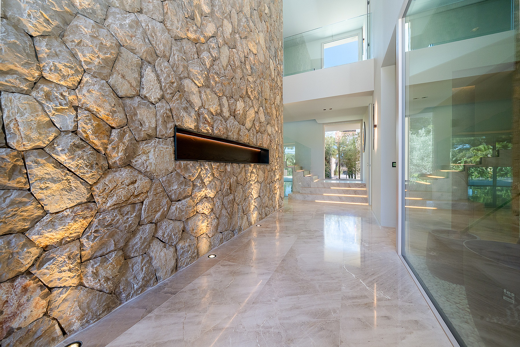 Interior entrance hall with a large dry-stone feature wall, linear fireplace, polished light marble floors, and glass partitions.
