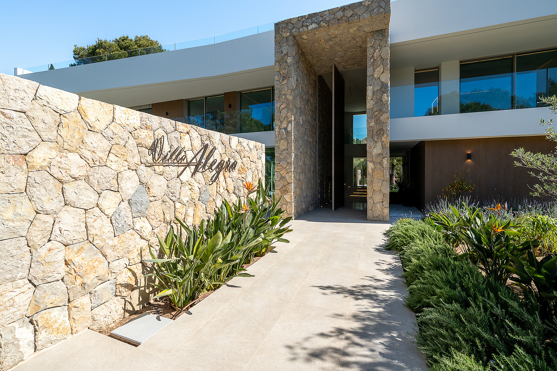 Modern villa entrance featuring a curved natural stone retaining wall, paved walkway, and landscaping in Santa Ponsa.