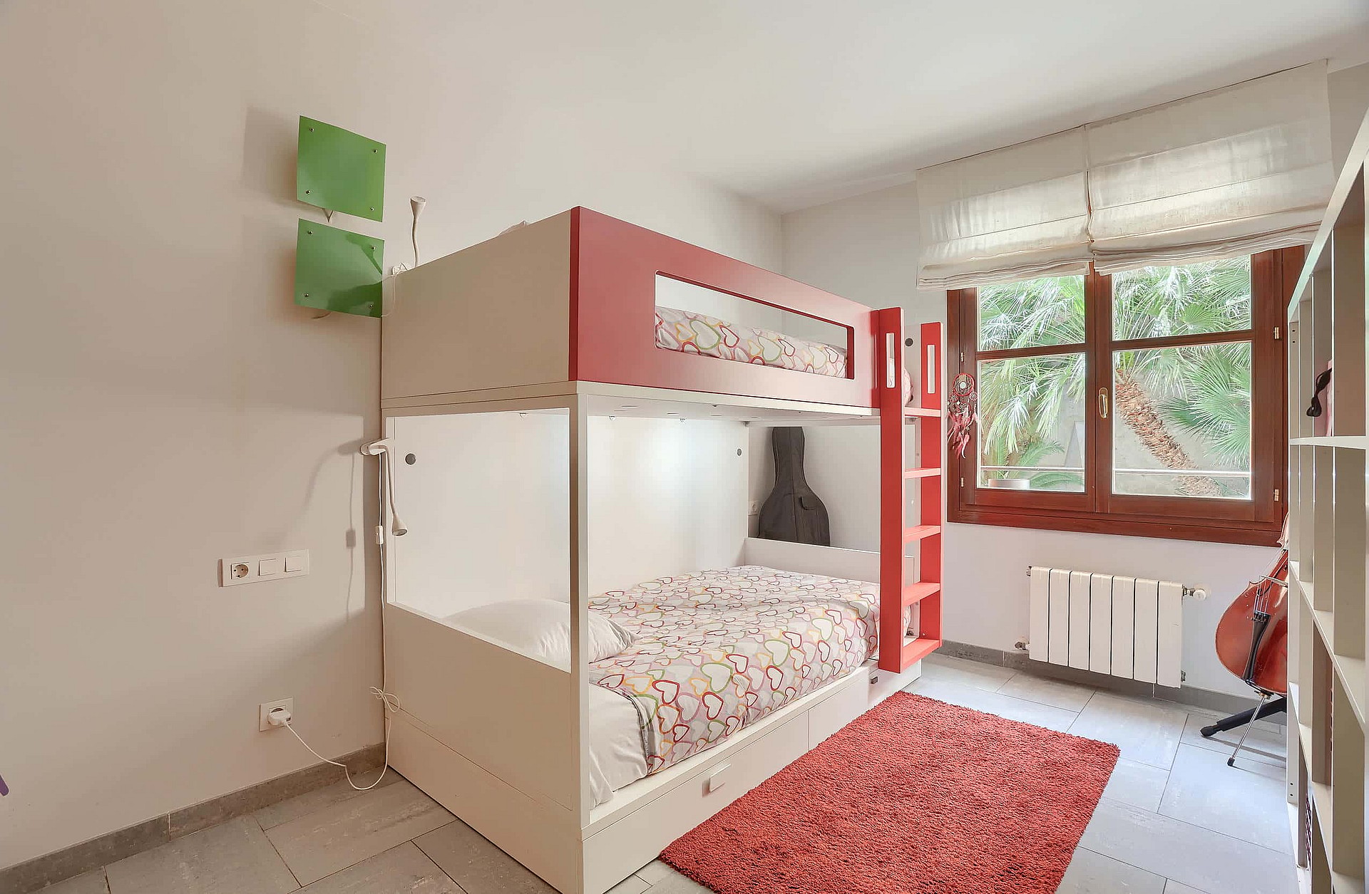 A bedroom featuring a red and white bunk bed, a red area rug, and a window with white Roman blinds.
