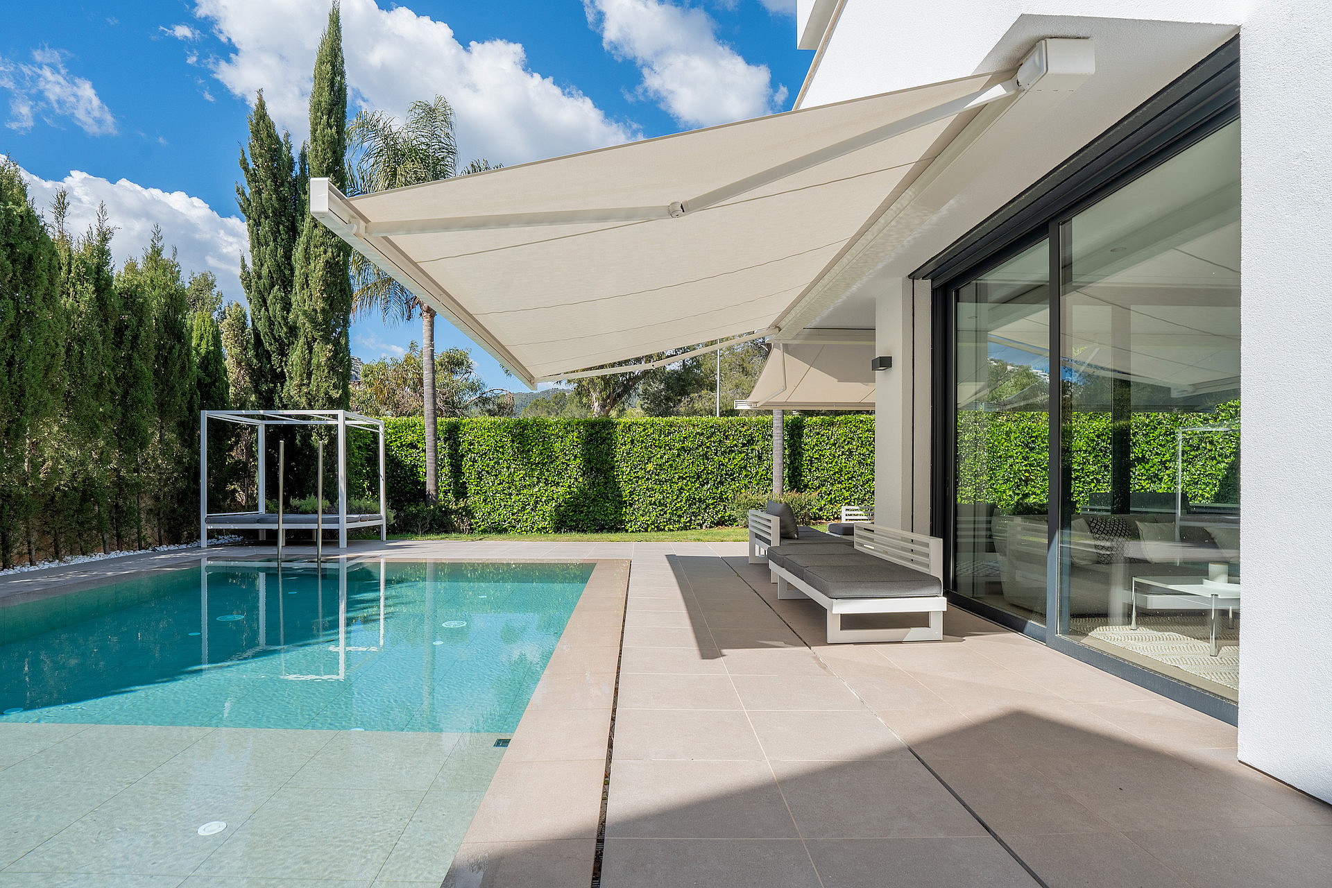 Heated saltwater swimming pool with sun loungers, retractable awning and tall hedge screening.