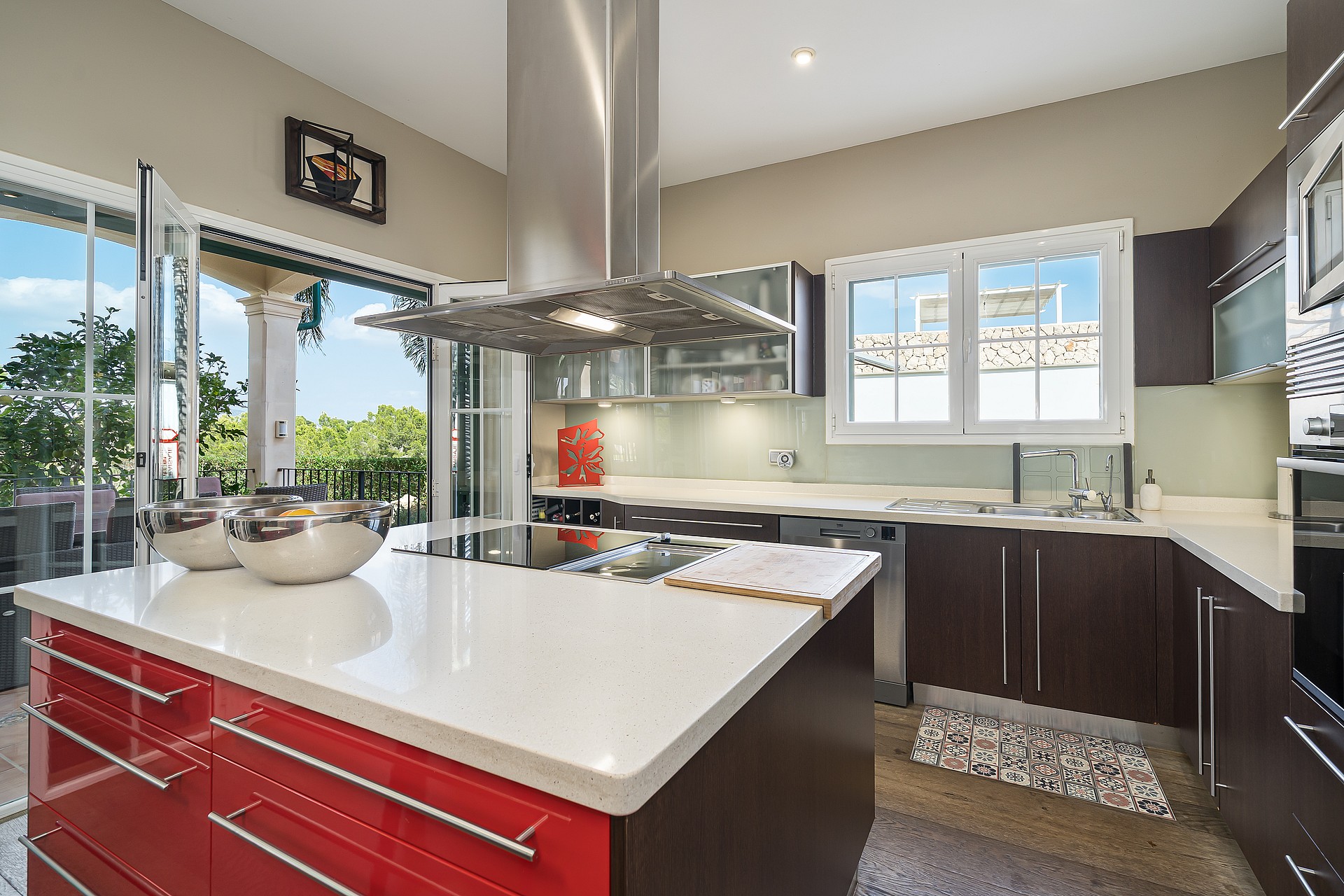 A modern kitchen featuring a bold red central island with a white stone countertop, dark cabinetry, and floor-to-ceiling glass doors opening to a terrace.