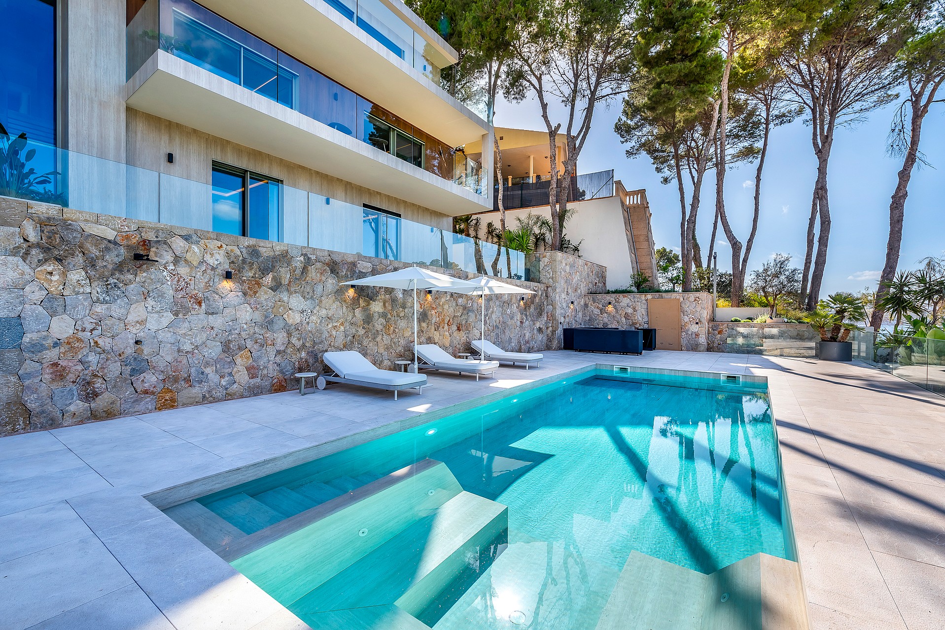 Outdoor swimming pool with white loungers on a paved terrace, set against stone walls and pine trees under a clear blue sky.