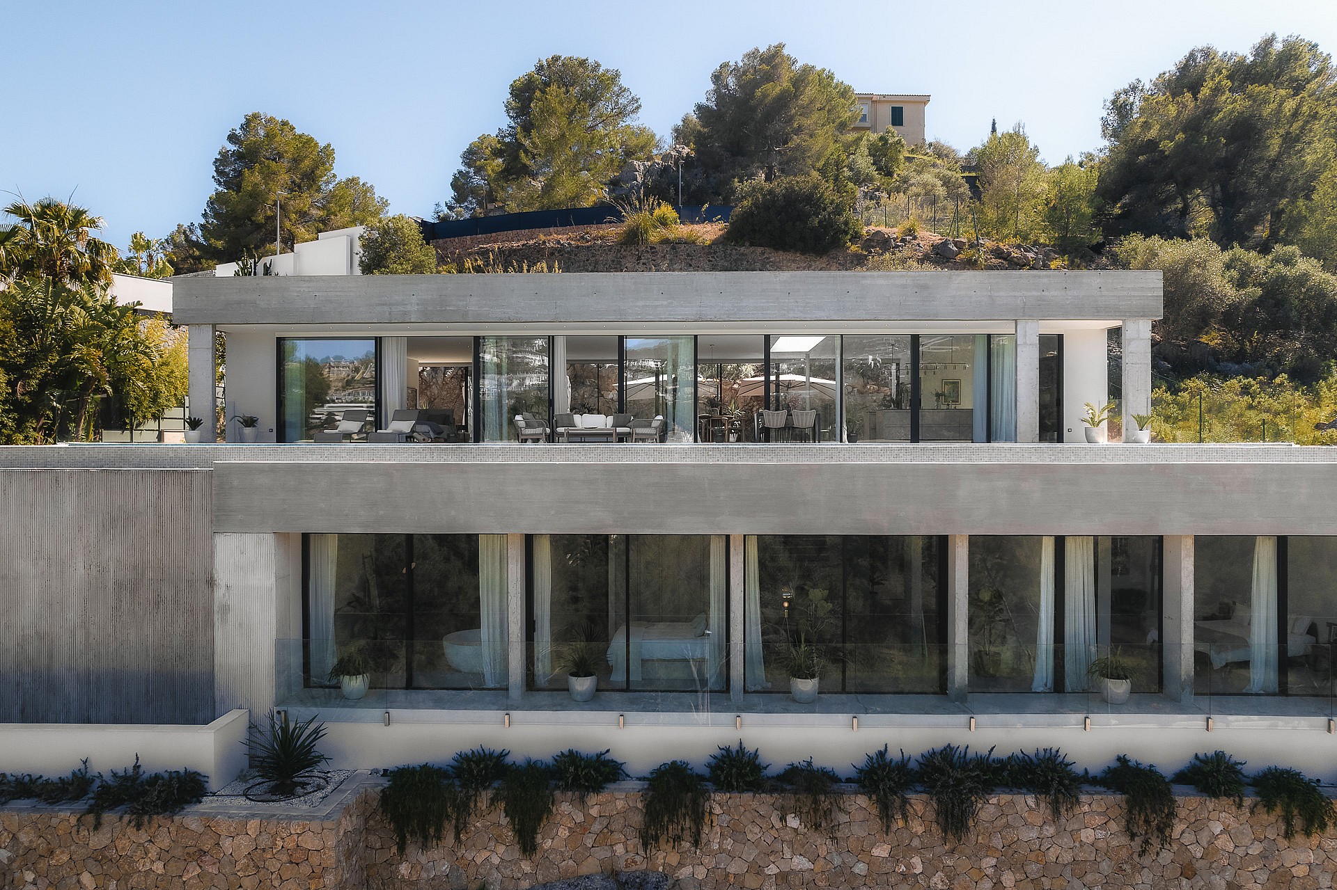 A two-level contemporary villa with exposed concrete, floor-to-ceiling glass, and stone walls on a Mediterranean hillside.