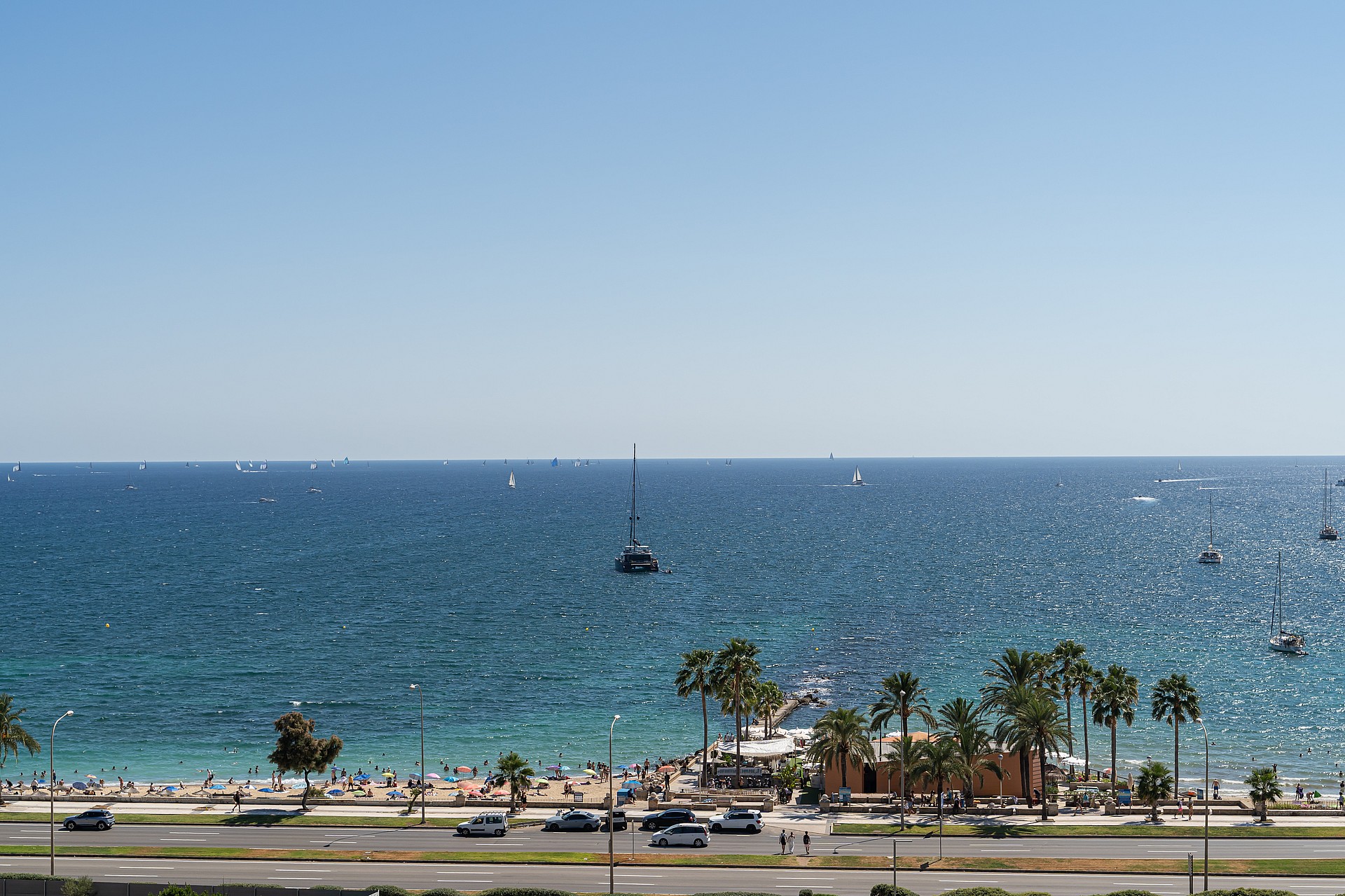 Wide angle view of the sparkling sea and coastline from an elevated balcony, featuring a sandy beach and palm trees.