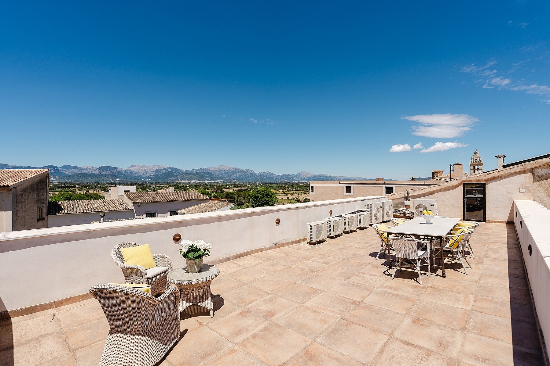 Wide rooftop terrace with terracotta floor tiles, wicker seating, and a dining set, looking out towards distant mountains.