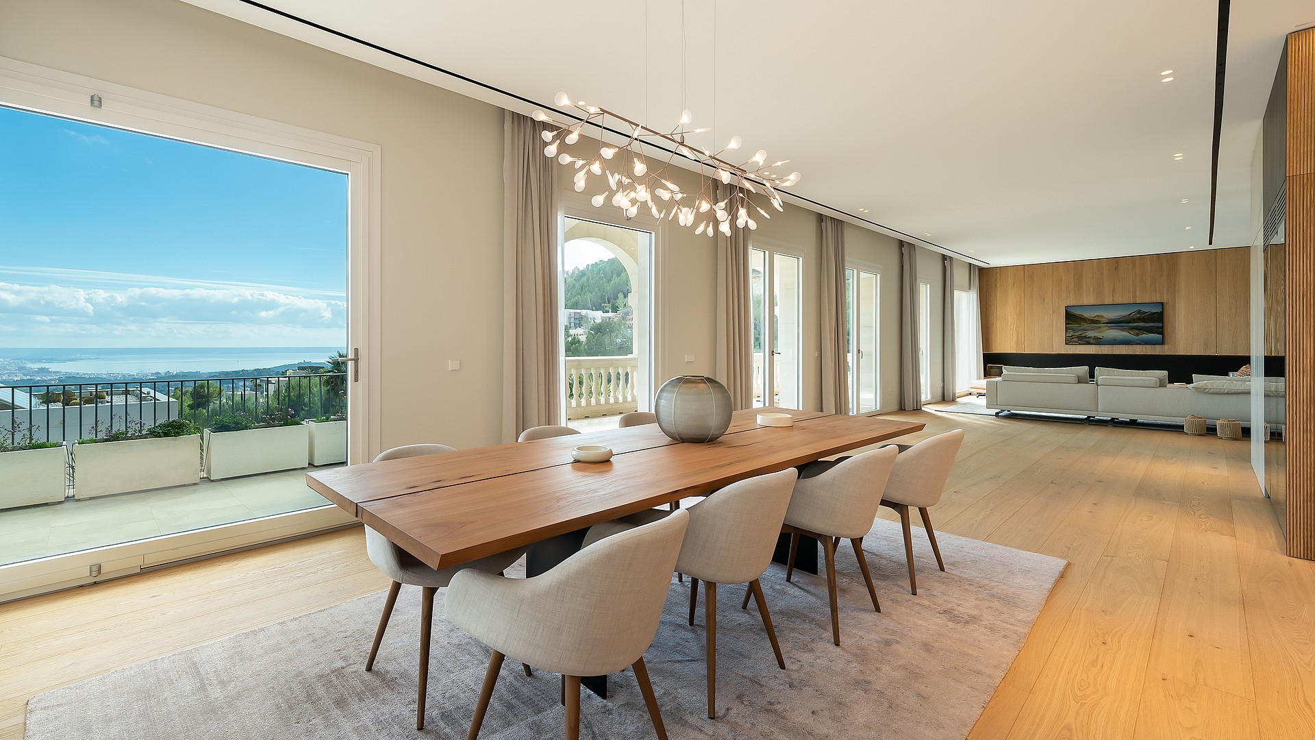Dining room featuring a long wooden table, light-colored upholstered chairs, and a sculptural chandelier, overlooking the sea.