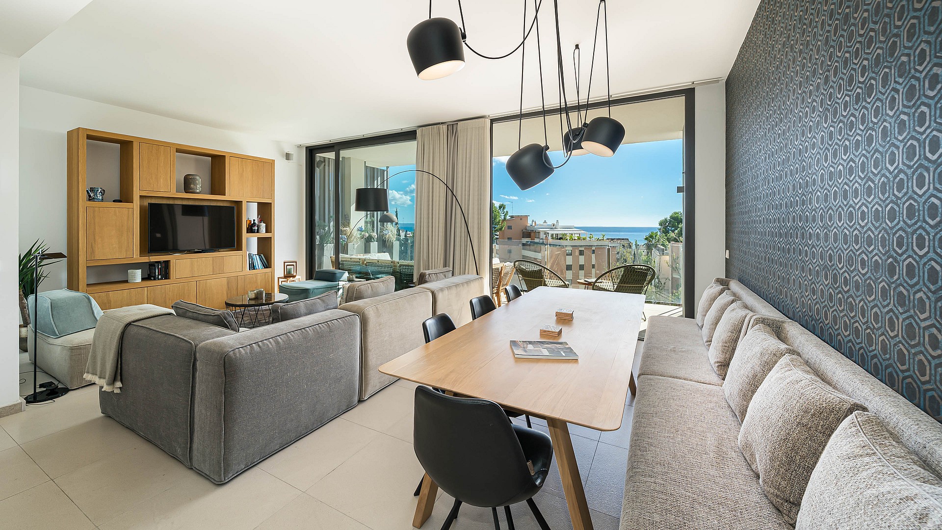 A modern open-plan living room with a large grey sofa, a wooden dining table, and floor-to-ceiling glass doors opening to a terrace with ocean views.