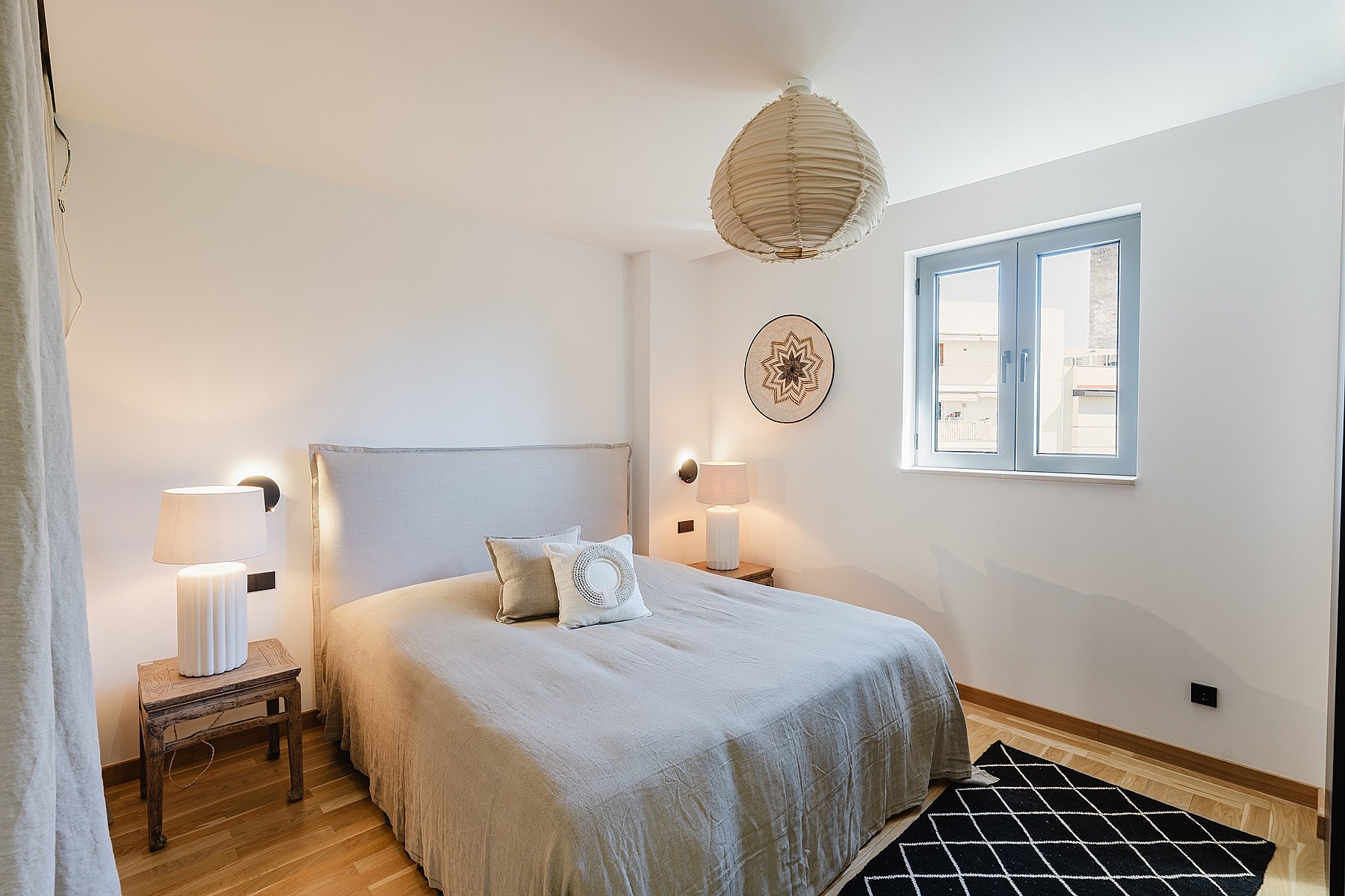 A bright bedroom with a light grey upholstered headboard, beige bedding, a paper pendant lamp, and a geometric patterned rug.